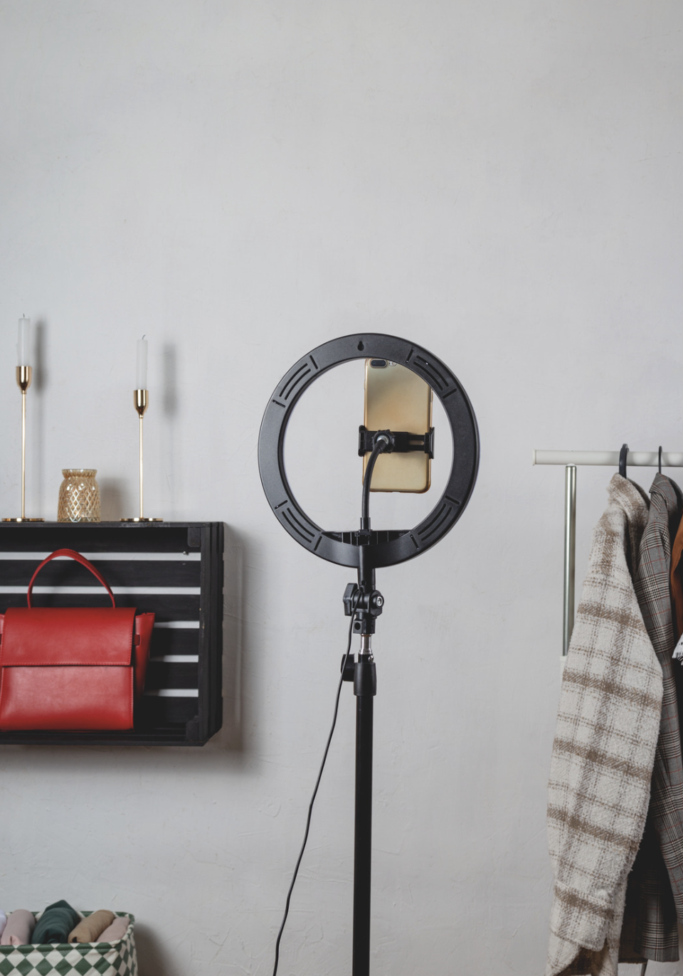 A ring light with a smartphone holder and a phone attached, set up indoors with a wall-mounted shelf holding candles and a red handbag, next to a clothing rack with plaid and beige coats.