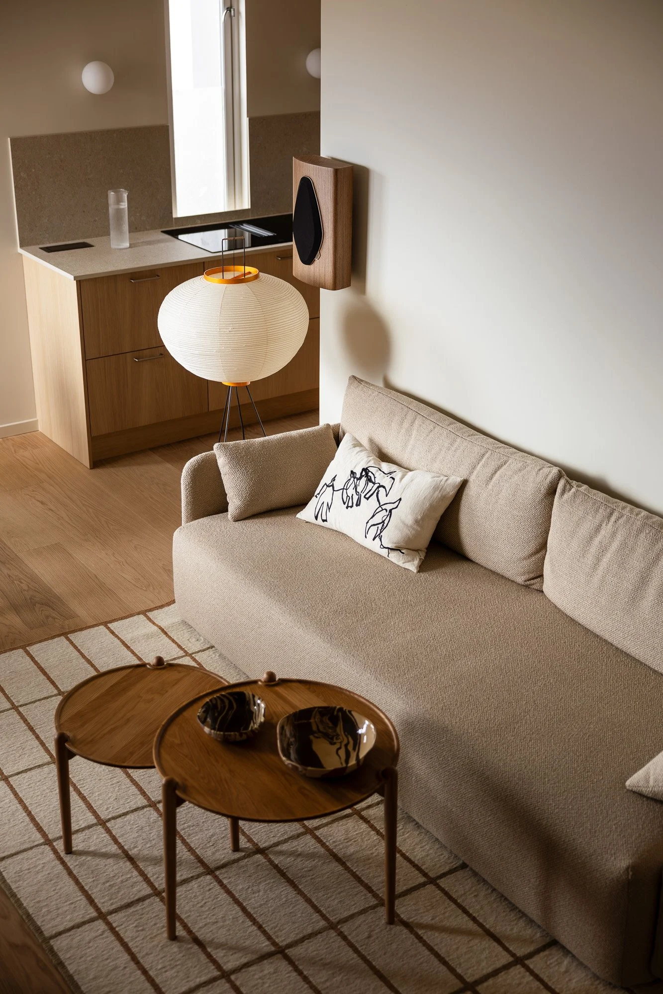 A living room with a beige sofa, a white pillow with black line art, two wooden coffee tables with marble bowls, a patterned rug, a wall-mounted speaker, a floor lamp with a round paper shade, and a small kitchen area with wood cabinets, a window, and a sink.