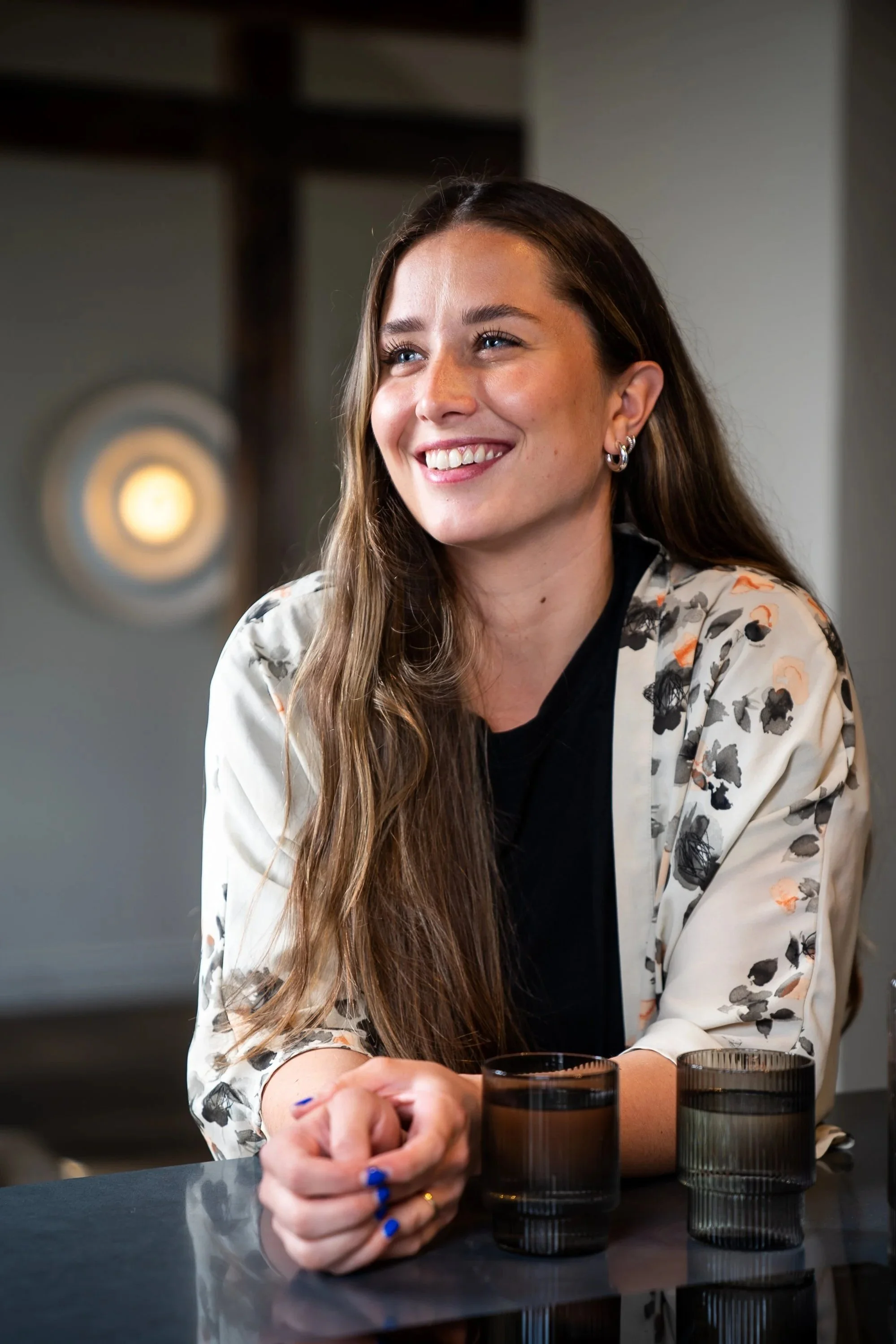 A smiling woman with long brown hair, wearing earrings and a floral blazer, sitting at a dark table with two glasses of water, in a modern indoor setting.