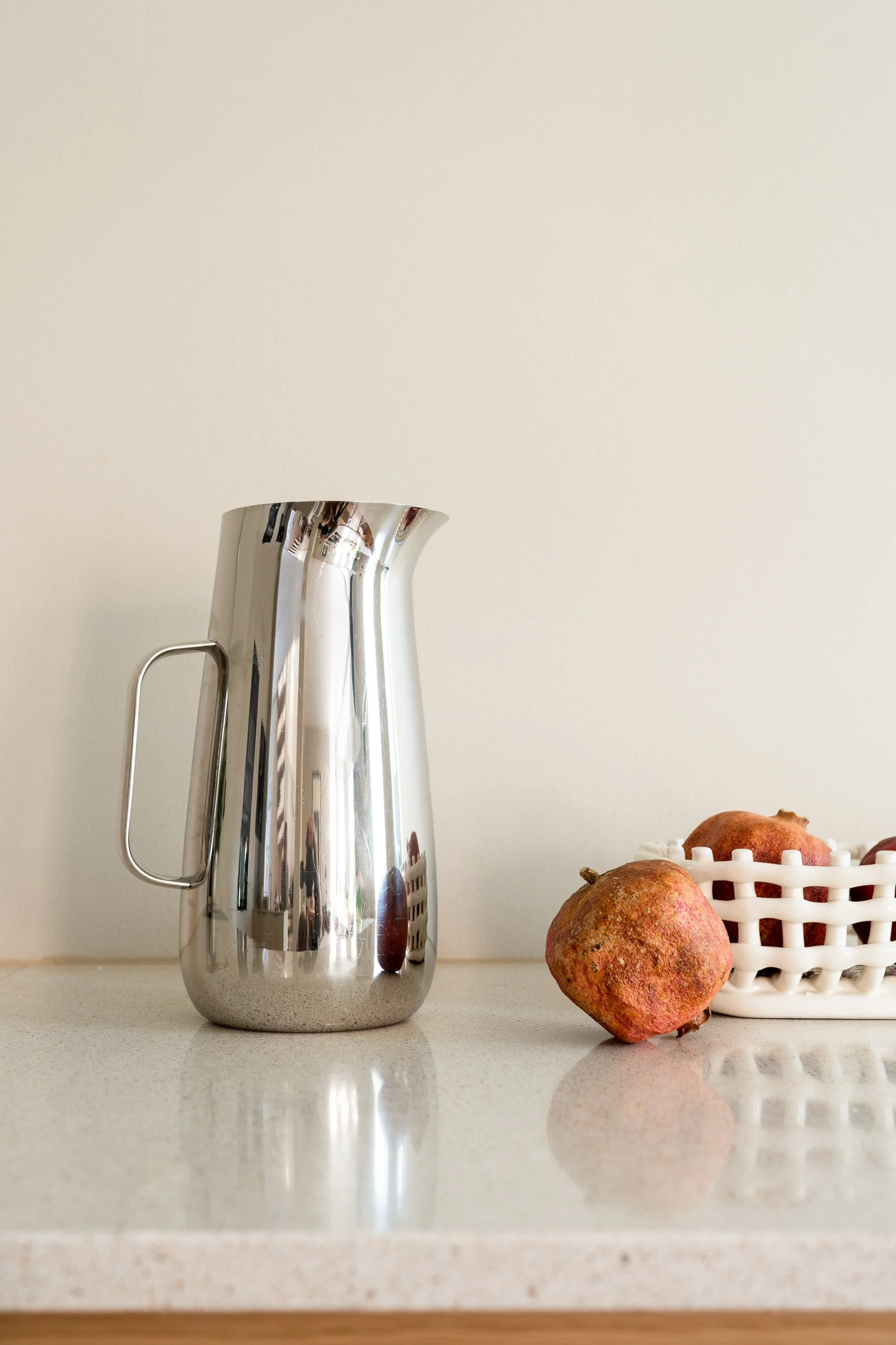 A reflective metal pitcher and a basket of pomegranates on a white countertop against a plain wall.