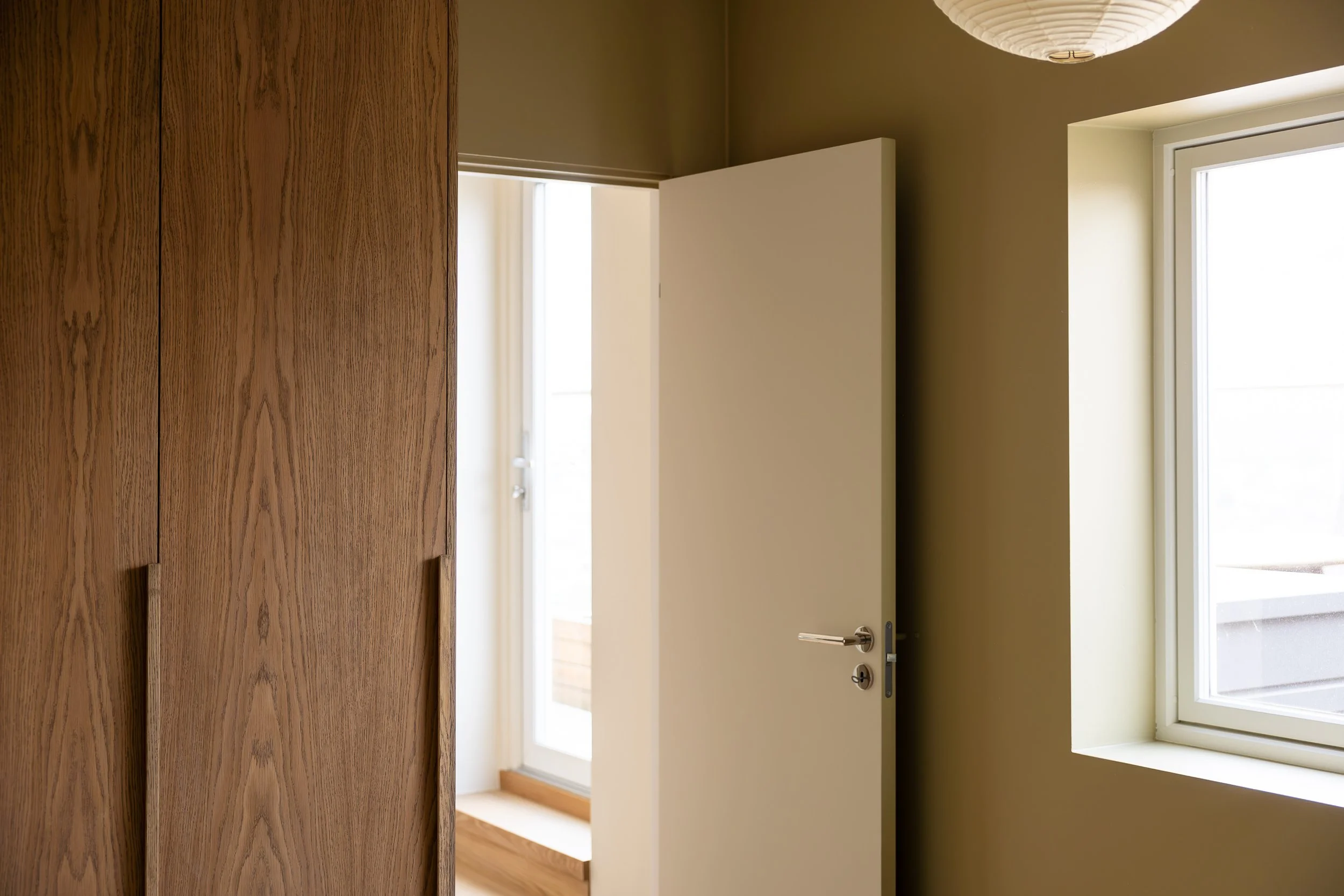Interior view of a room with a wooden wardrobe, an open white door, and a window letting in natural light.