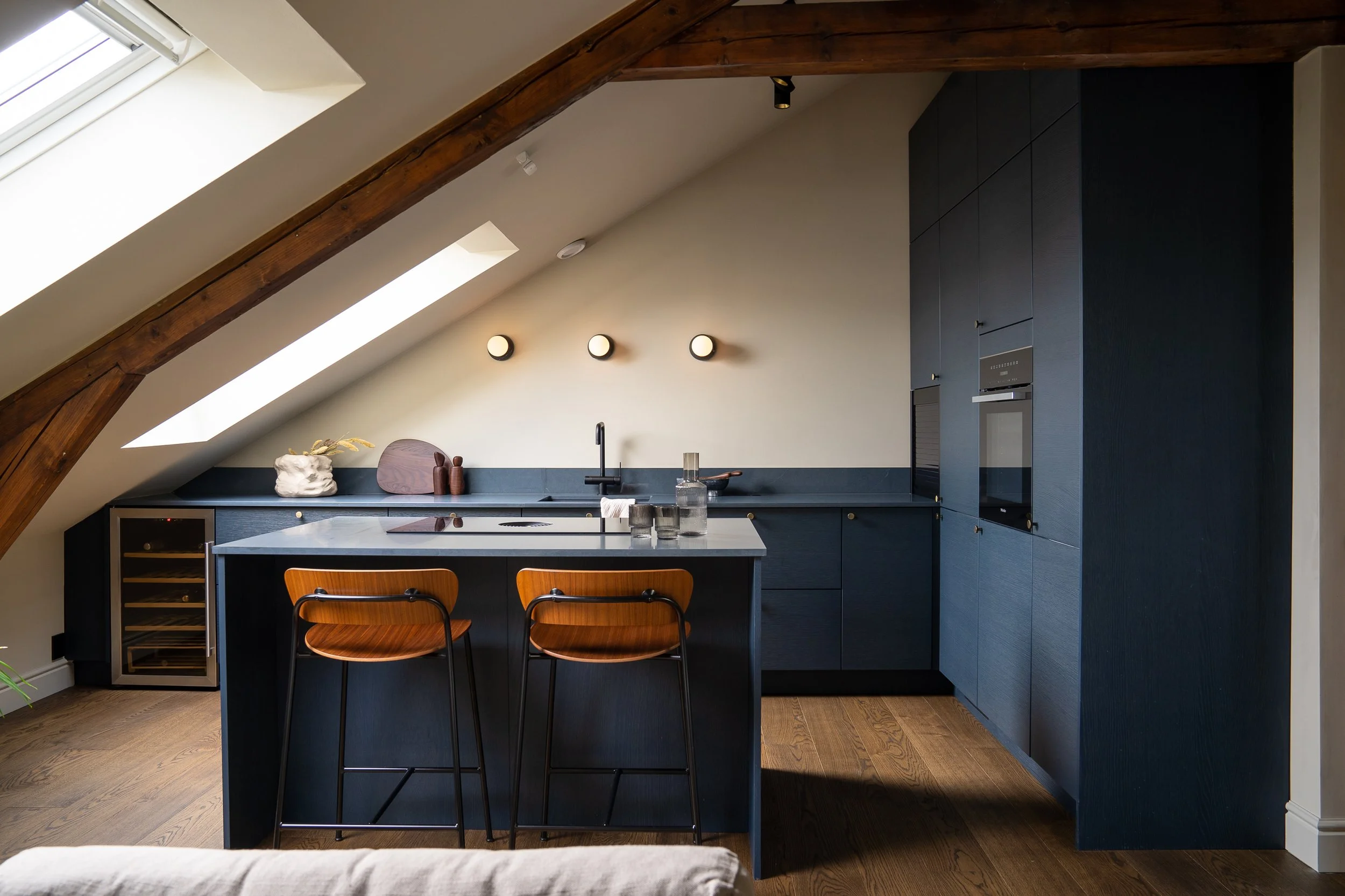 Modern attic kitchen with blue cabinets, wooden ceiling beams, skylight windows, and a kitchen island with two wooden bar stools.
