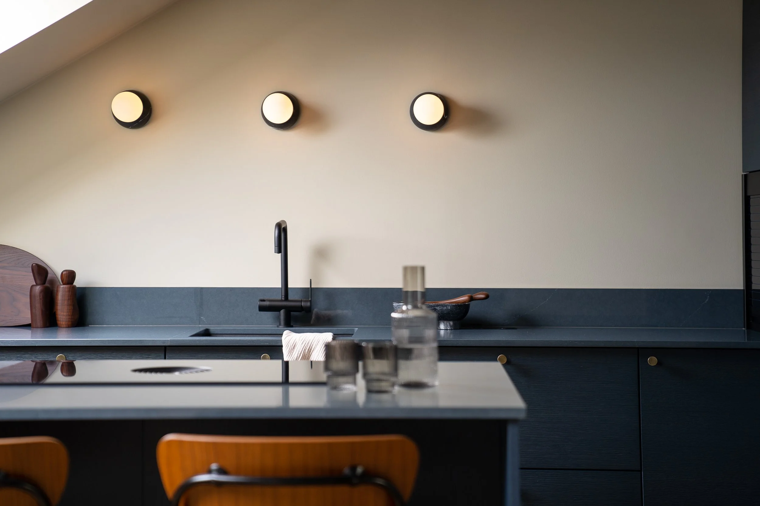 Modern kitchen with dark cabinets, black sink and faucet, beige wall with three round wall lights, and a countertop with glass jars and a wooden spoon.