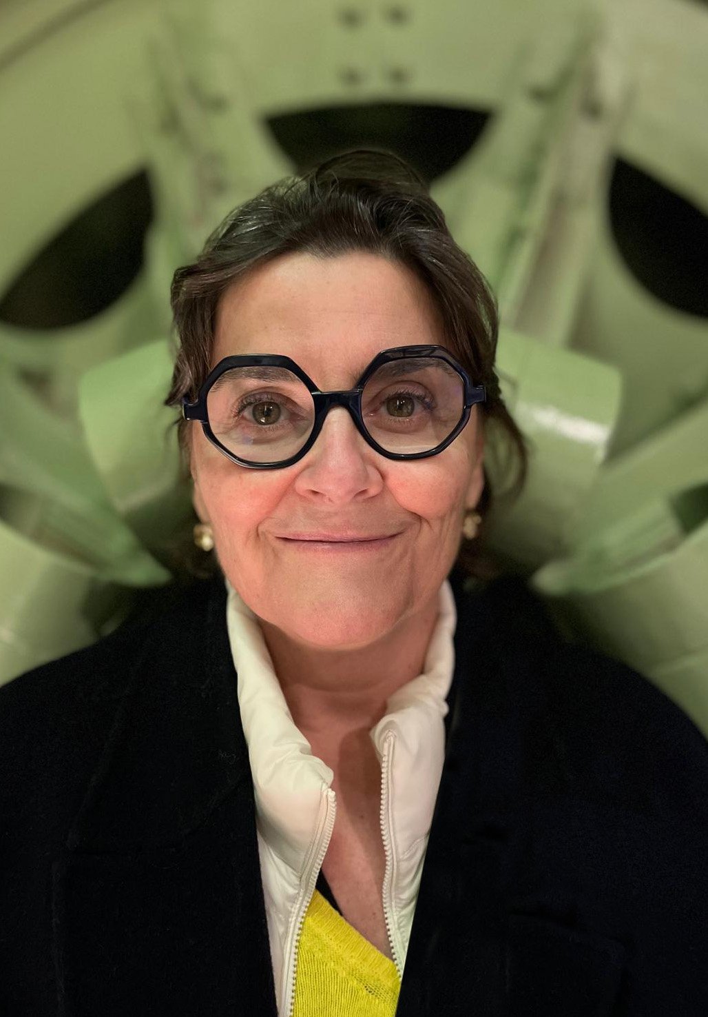 Une femme souriante portant des lunettes rondes noires, une veste noire, une chemise blanche et un pull jaune, avec un fond de plantes vertes.