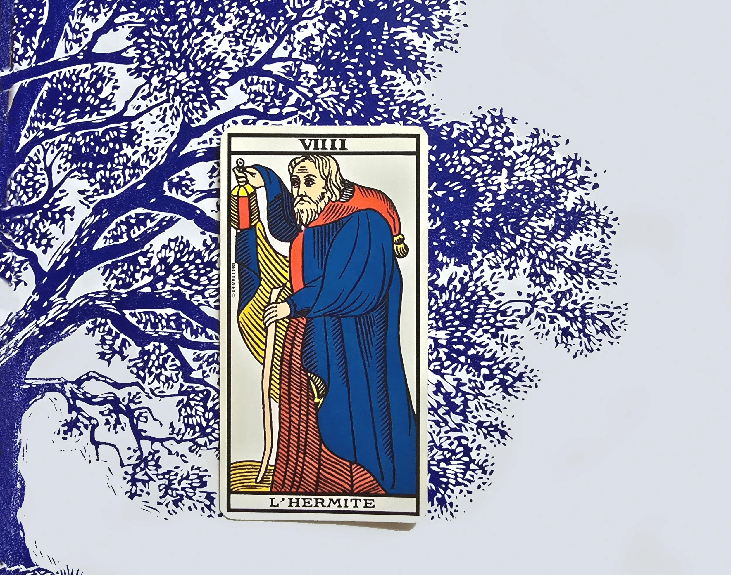 Une carte de tarot intitulée L'Hermite, représentant un homme âgé avec une longue barbe, portant une tunique bleue et un manteau rouge, tenant une lanterne dans sa main gauche et un bâton dans sa main droite, avec un fond d'arbre stylisé en bleu.