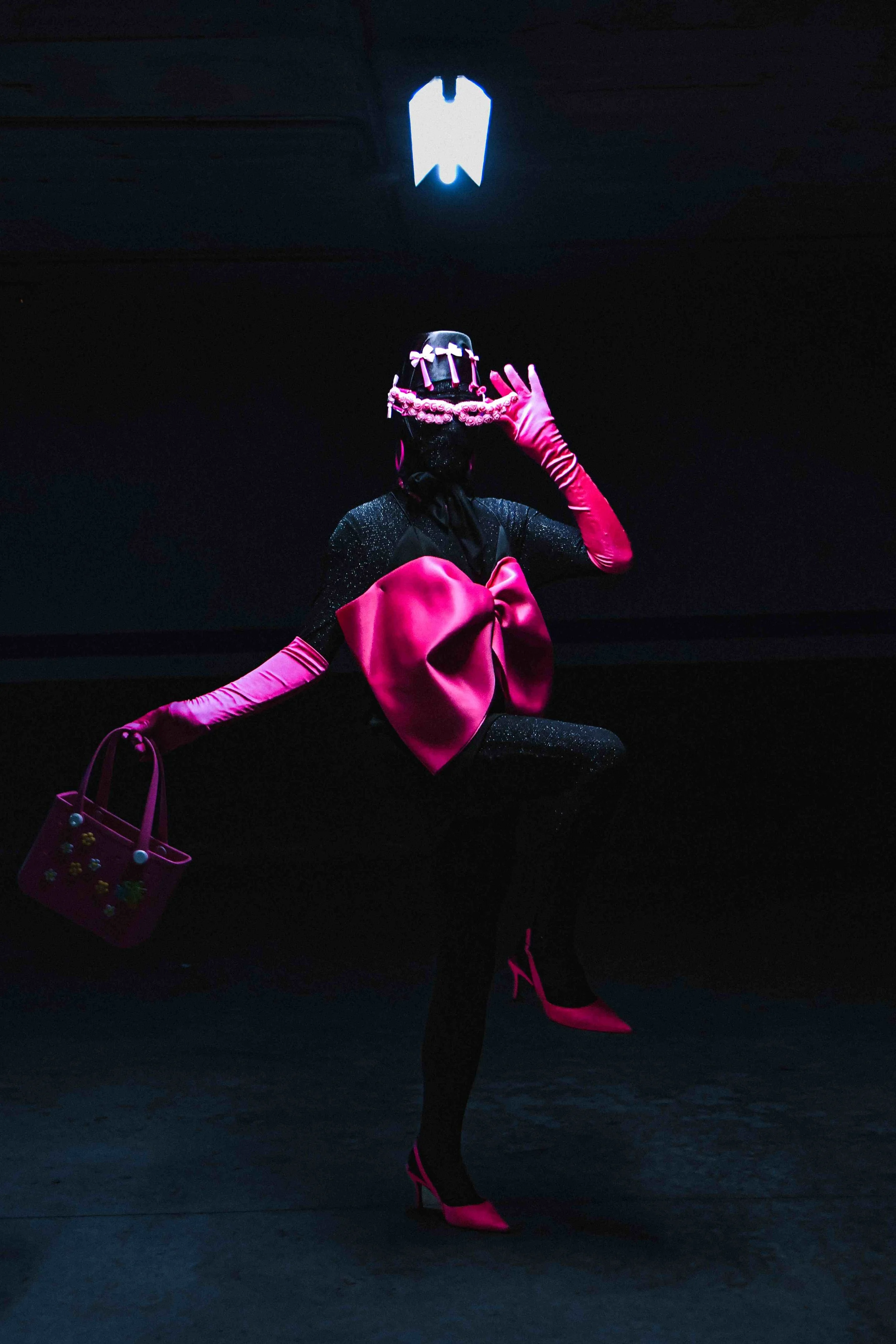 Salma Zahore, a queer Arab icon, wears an all-black catsuit with a large pink bow, matching pink gloves, and shoes, standing in the middle of a dark parking lot.