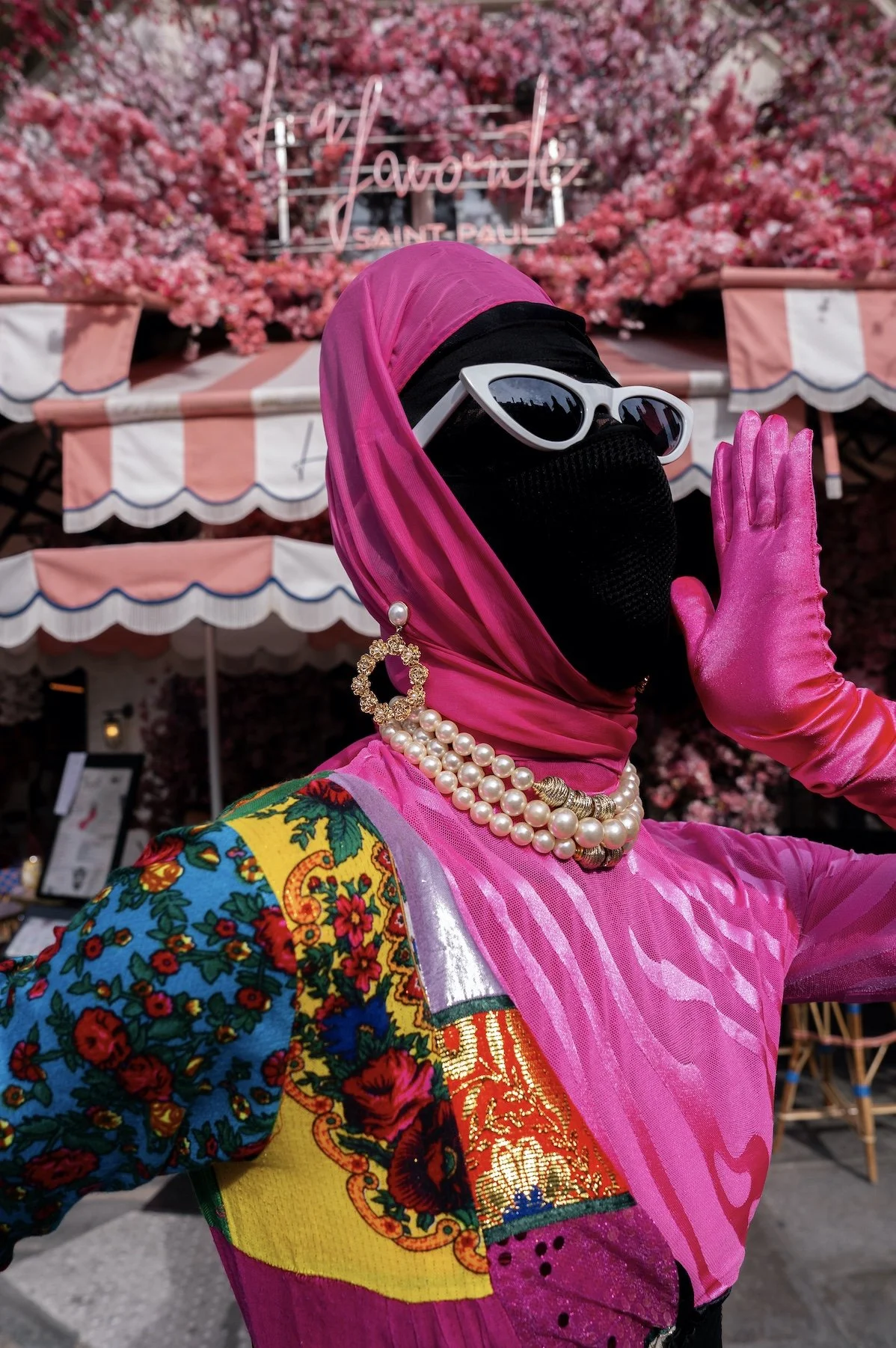 Salma Zahore dressed in colorful abaya, floral-patterned clothing and pink accessories, wearing a pink headscarf, white sunglasses, pink gloves, and pearl jewelry, standing in front of a pink floral backdrop.