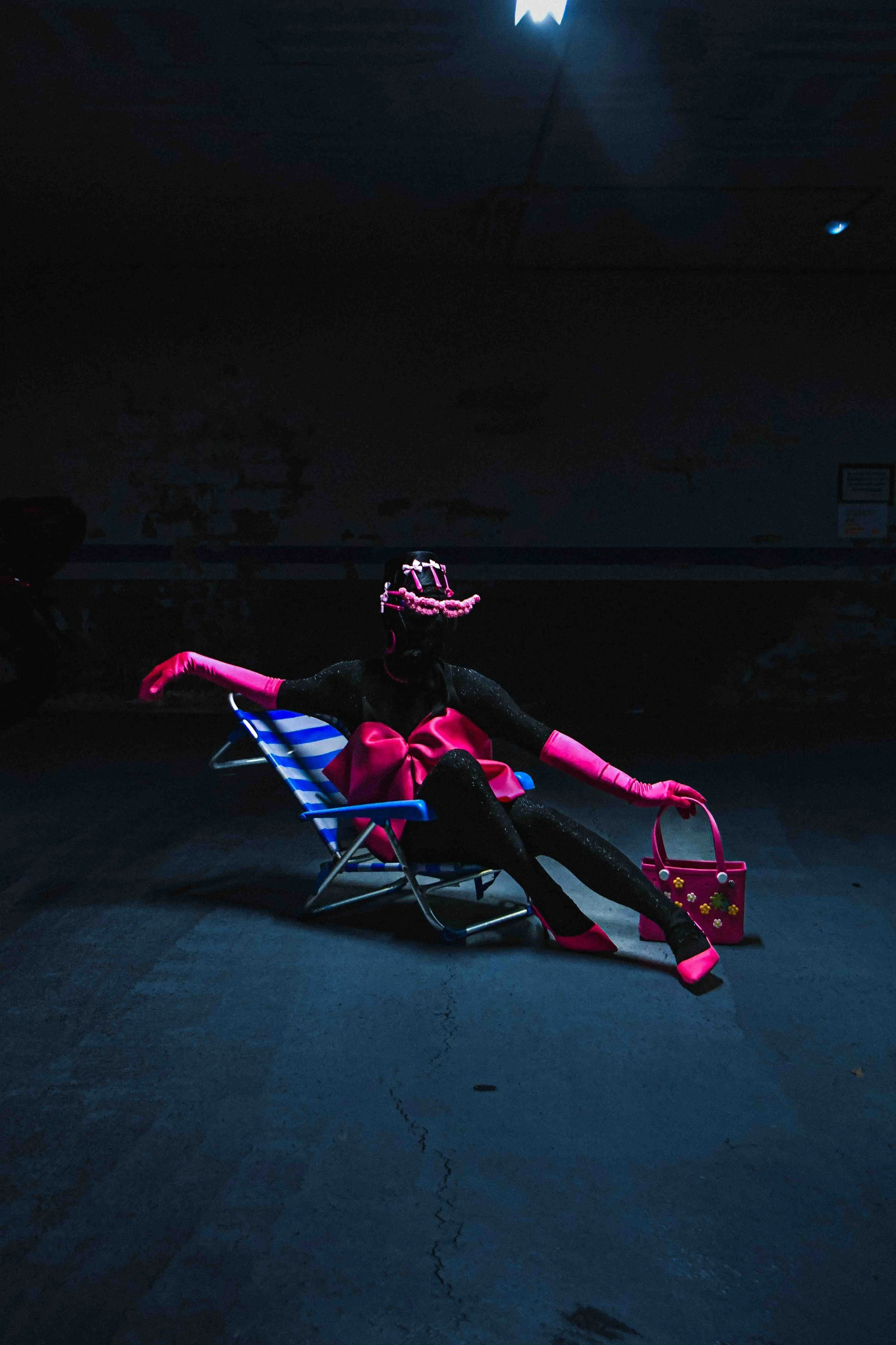 Salma Zahore, a queer Arab icon, wears an all-black catsuit with a large pink bow, matching pink gloves and shoes, standing in the middle of a dark parking lot. She is sitting on a chair. 