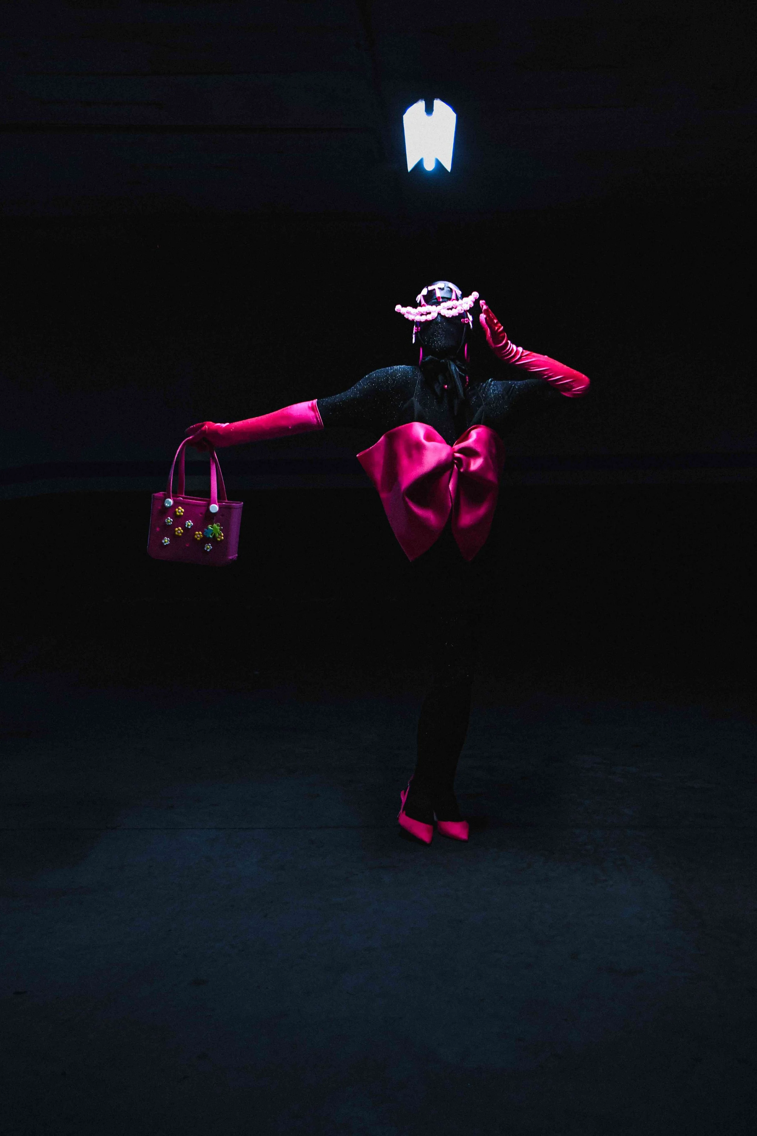 Salma Zahore, a queer Arab icon, wears an all-black catsuit with a large pink bow, matching pink gloves, and shoes, standing in the middle of a dark parking lot.