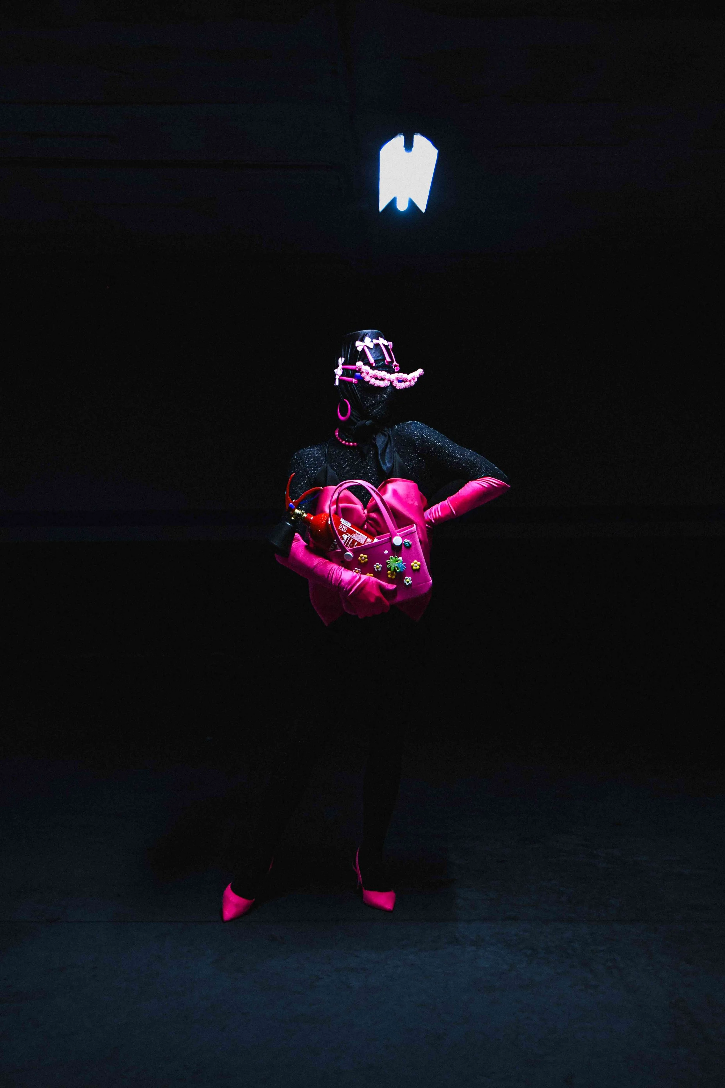 Salma Zahore, a queer Arab icon, wears an all-black catsuit with a large pink bow, matching pink gloves and shoes, standing in the middle of a dark parking lot. She holds her pink bag, adorned with the red Fire Instinctor.