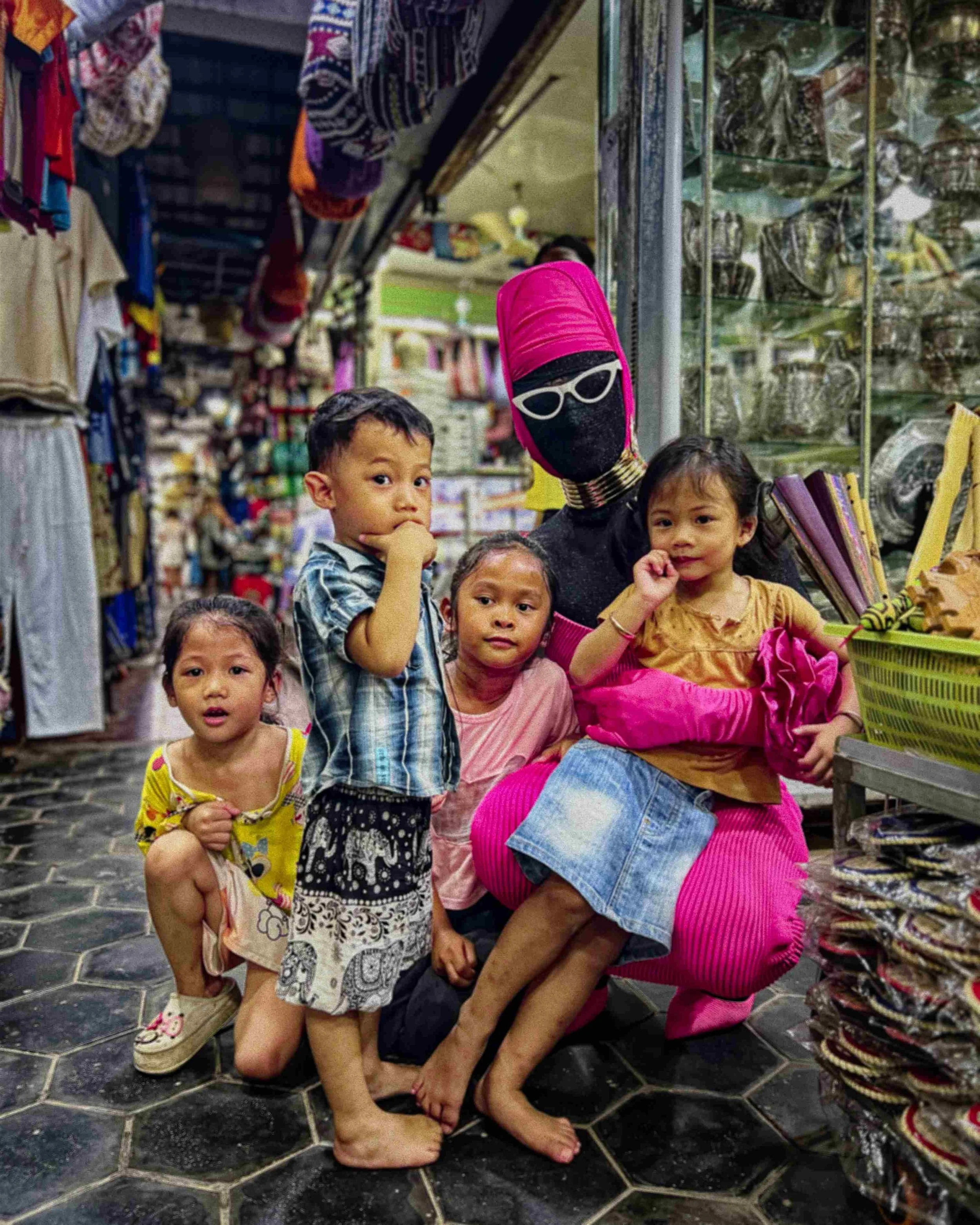 Salma Zahore, a queer Arab icon, wearing white glasses that cover her face, dressed in a full pink gown, in a supermarket in Siem Reap, Cambodia, with kids around her.