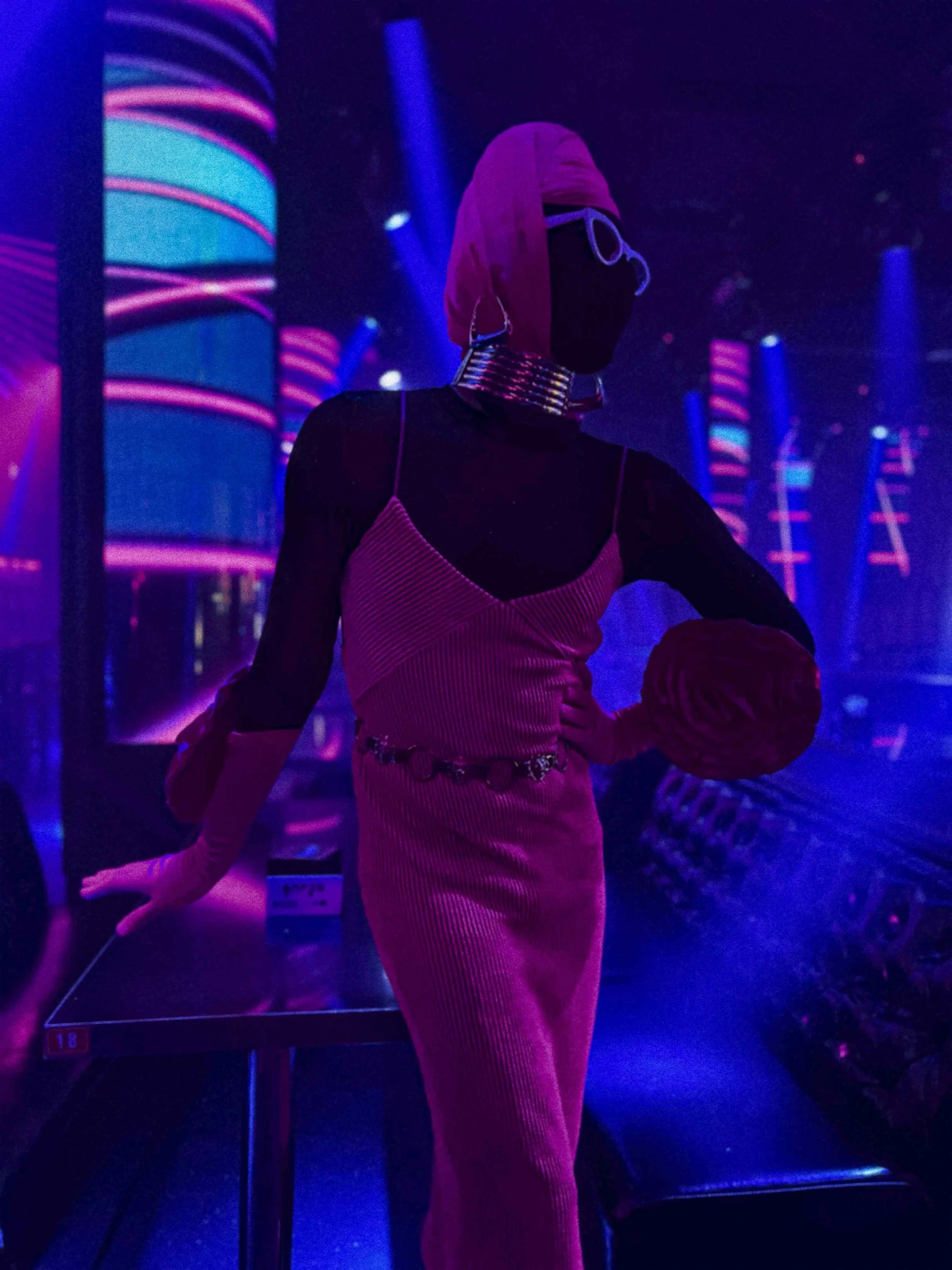 Salma Zahore, a queer Arab icon, wearing white glasses that cover her face, dressed in a full pink gown, in a nightclub in Siem Reap with a lot of neon lights around her.