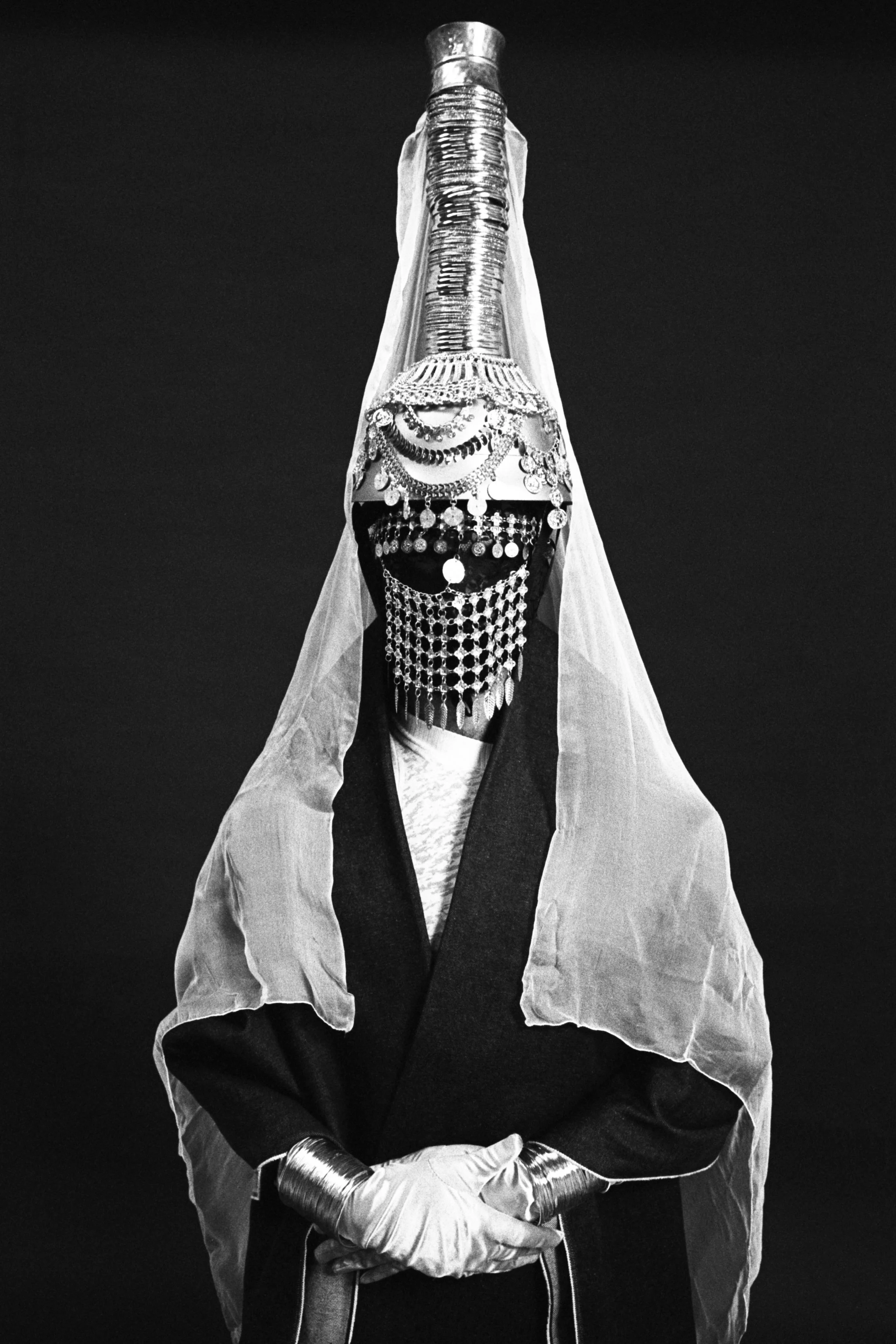 Salma Zahore is dressed in elaborate, layered costumes with metallic, beaded, and fabric accessories, with their face covered by a complex mask and jewelry. Salma Zahore is wearing a Tantour is standing with hands clasped against a dark background.