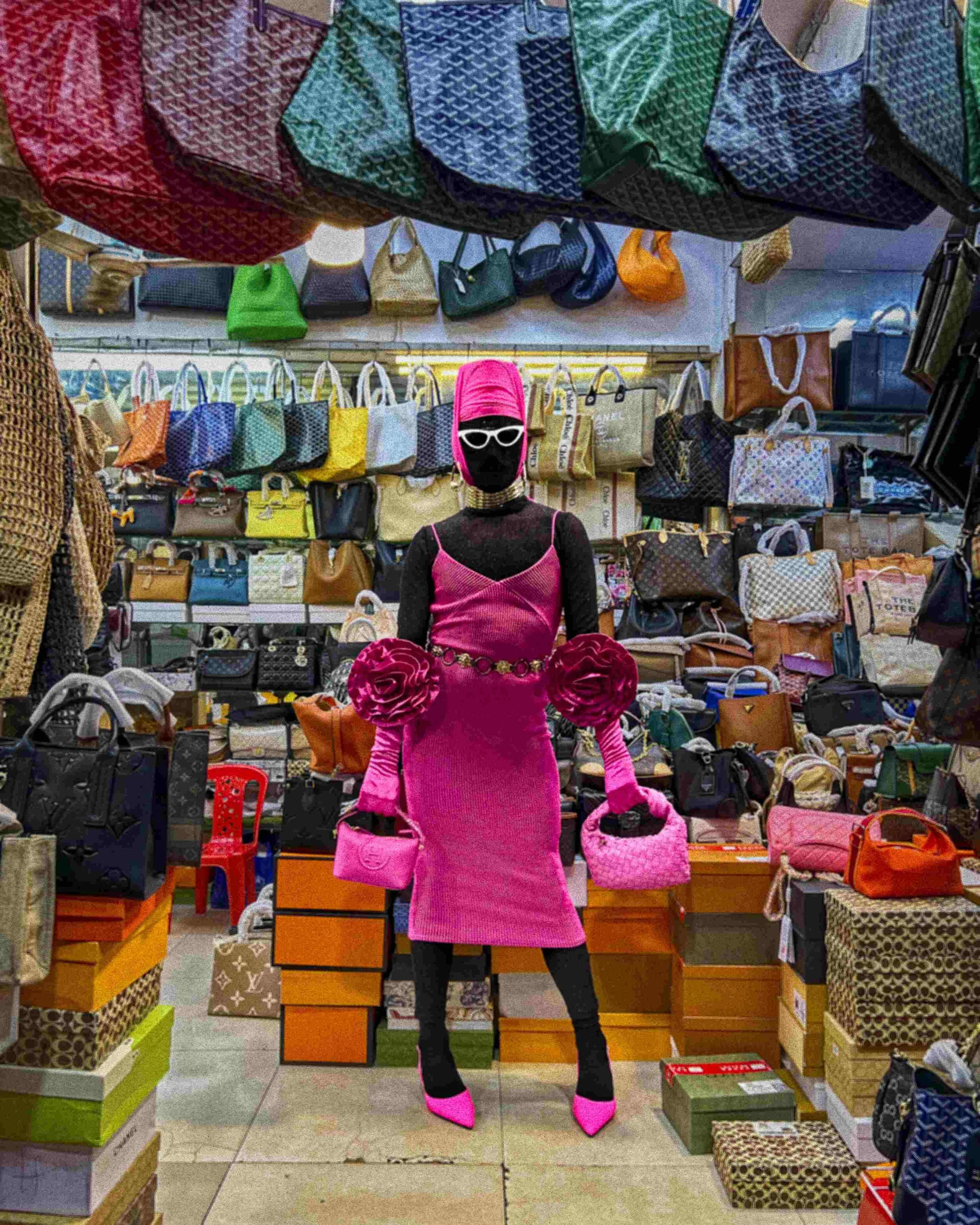 Salma Zahore, a queer Arab icon, wearing white glasses that cover her face, dressed in a full pink gown, in a supermarket in Siem Reap, Cambodia, holding two pink bags, one in each hand.
