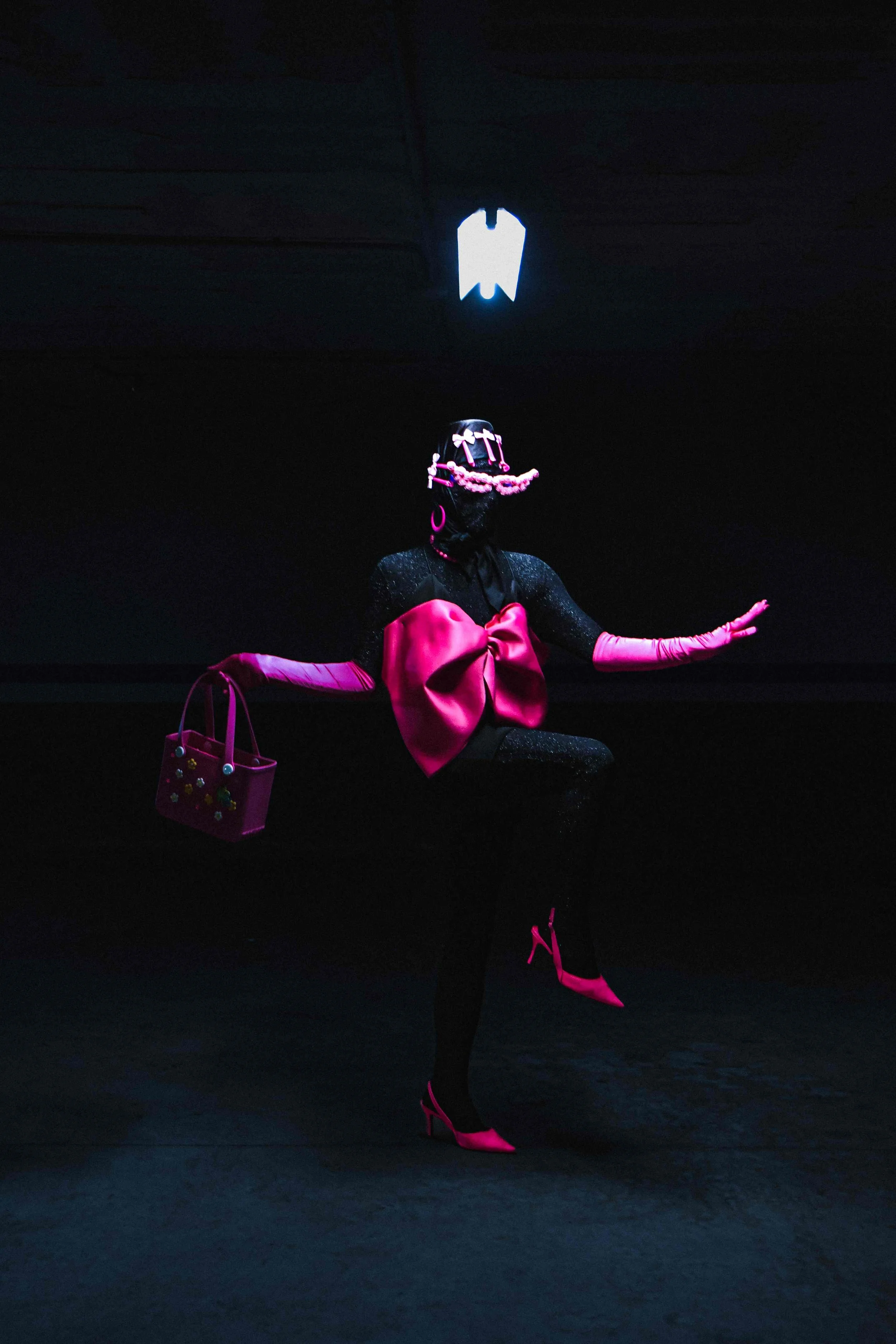 Salma Zahore, a queer Arab icon, wears an all-black catsuit with a large pink bow, matching pink gloves, and shoes, standing in the middle of a dark parking lot.