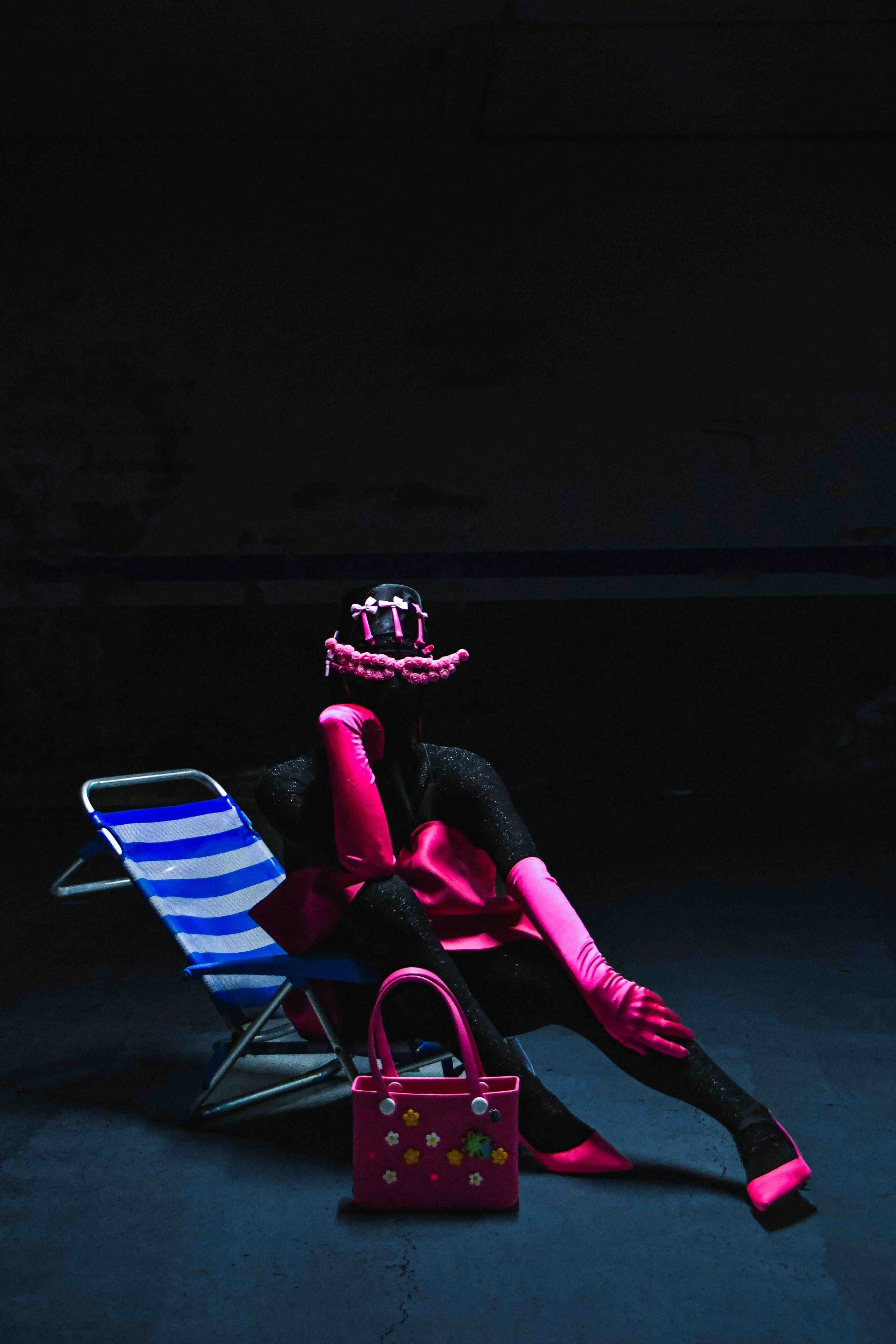 Salma Zahore, a queer Arab icon, wears an all-black catsuit with a large pink bow, matching pink gloves and shoes, standing in the middle of a dark parking lot. She is sitting on a chair. 
