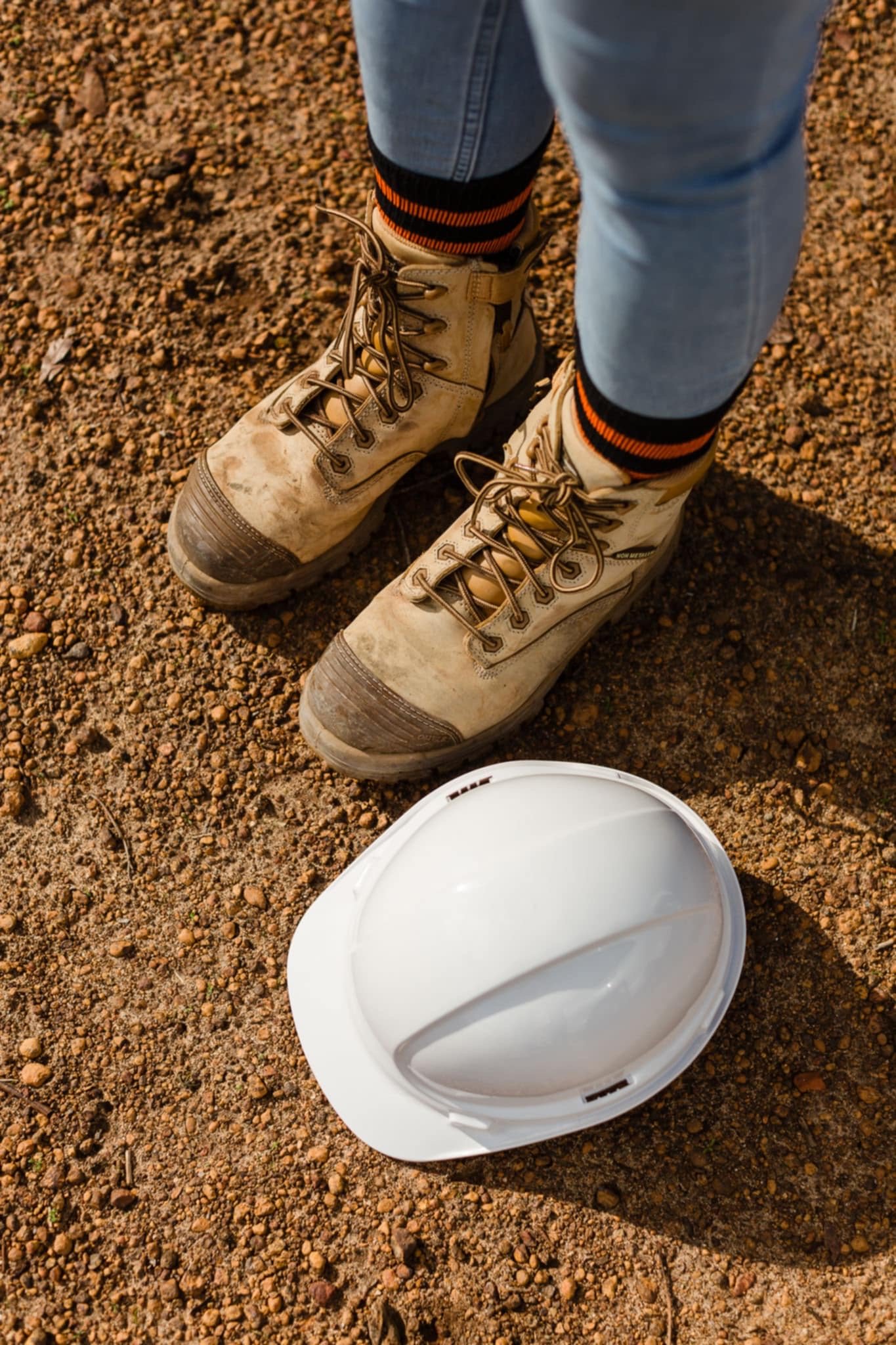A pair of tan work boots on dirt ground wearing blue jeans with rolled-up cuffs and striped orange and black socks. A white safety helmet is on the ground nearby.