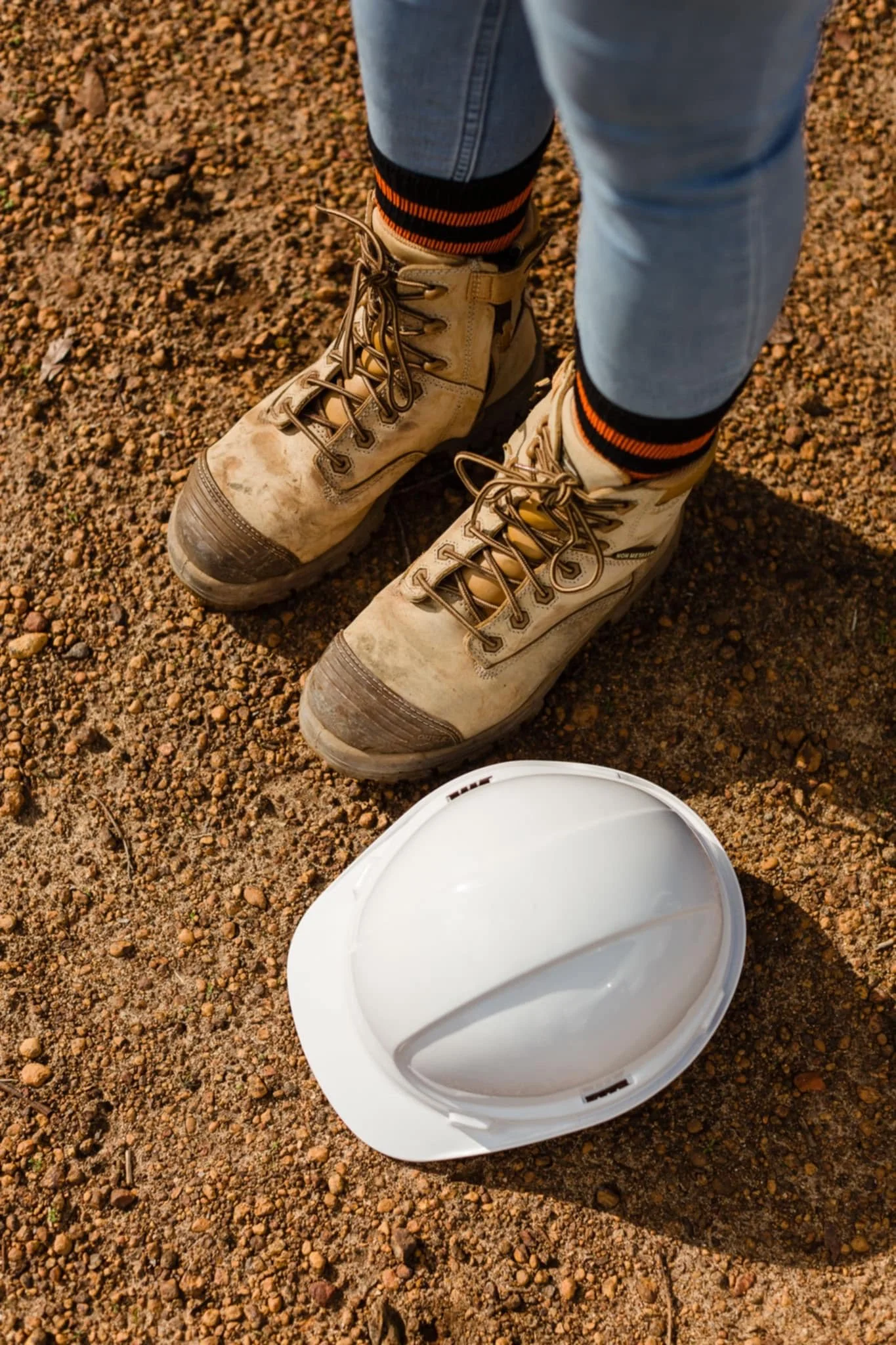 Close-up of a person wearing work boots standing on dirt, with a white safety helmet on the ground nearby.