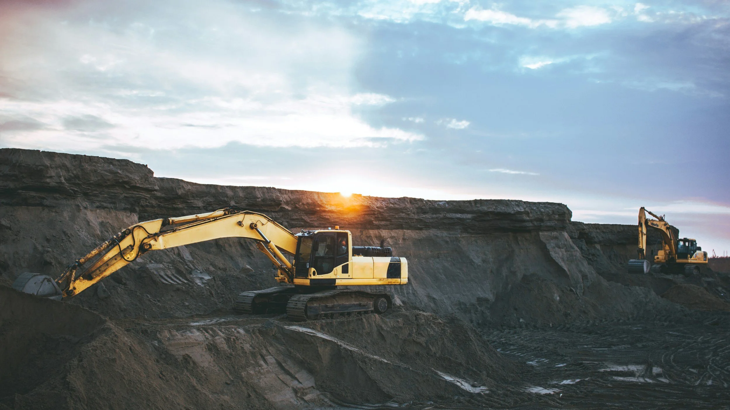 Two yellow excavators working on a dirt hill at sunset.