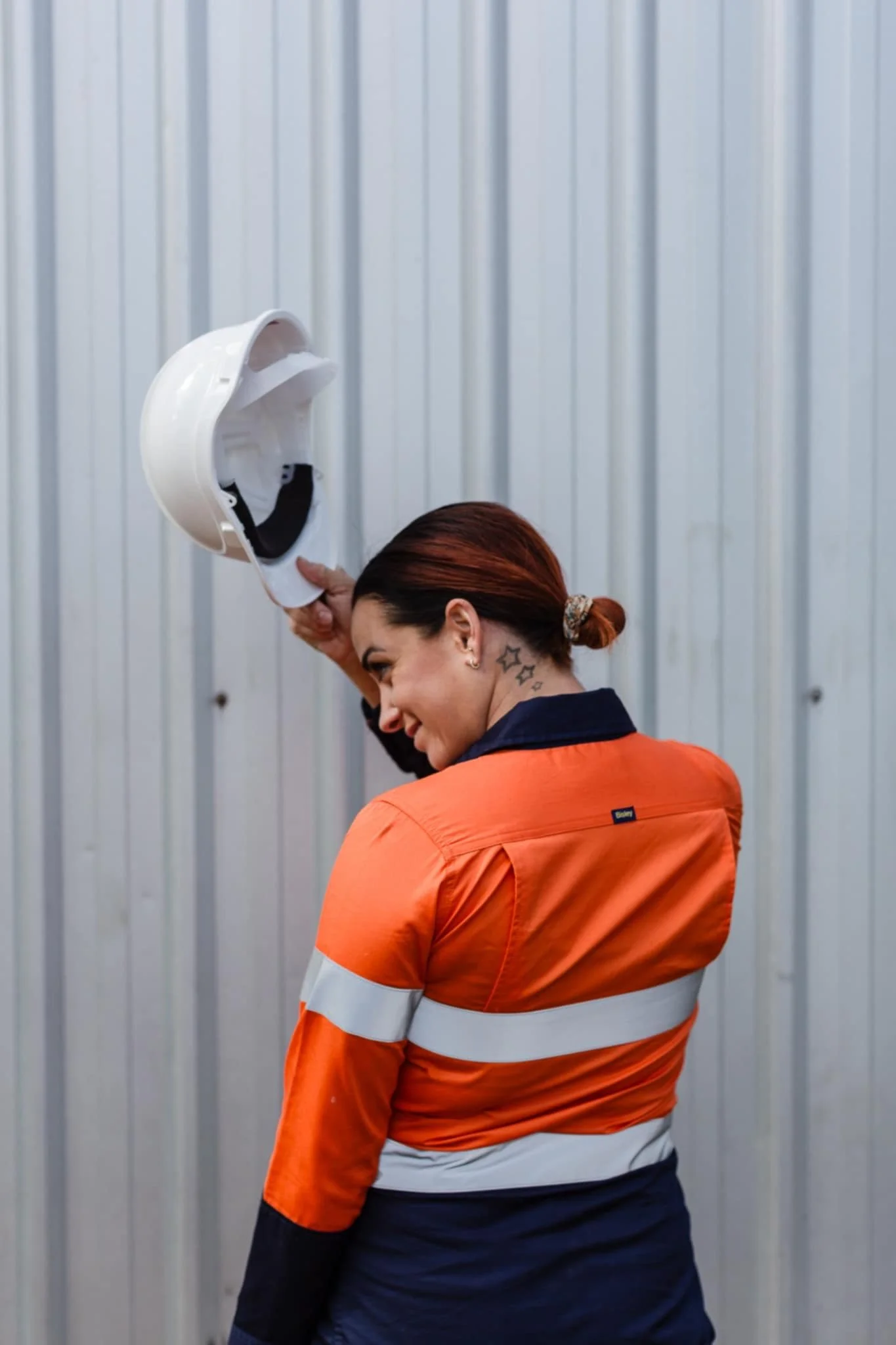 A woman in an orange safety jacket smiling and holding her hard hat against a metal wall.