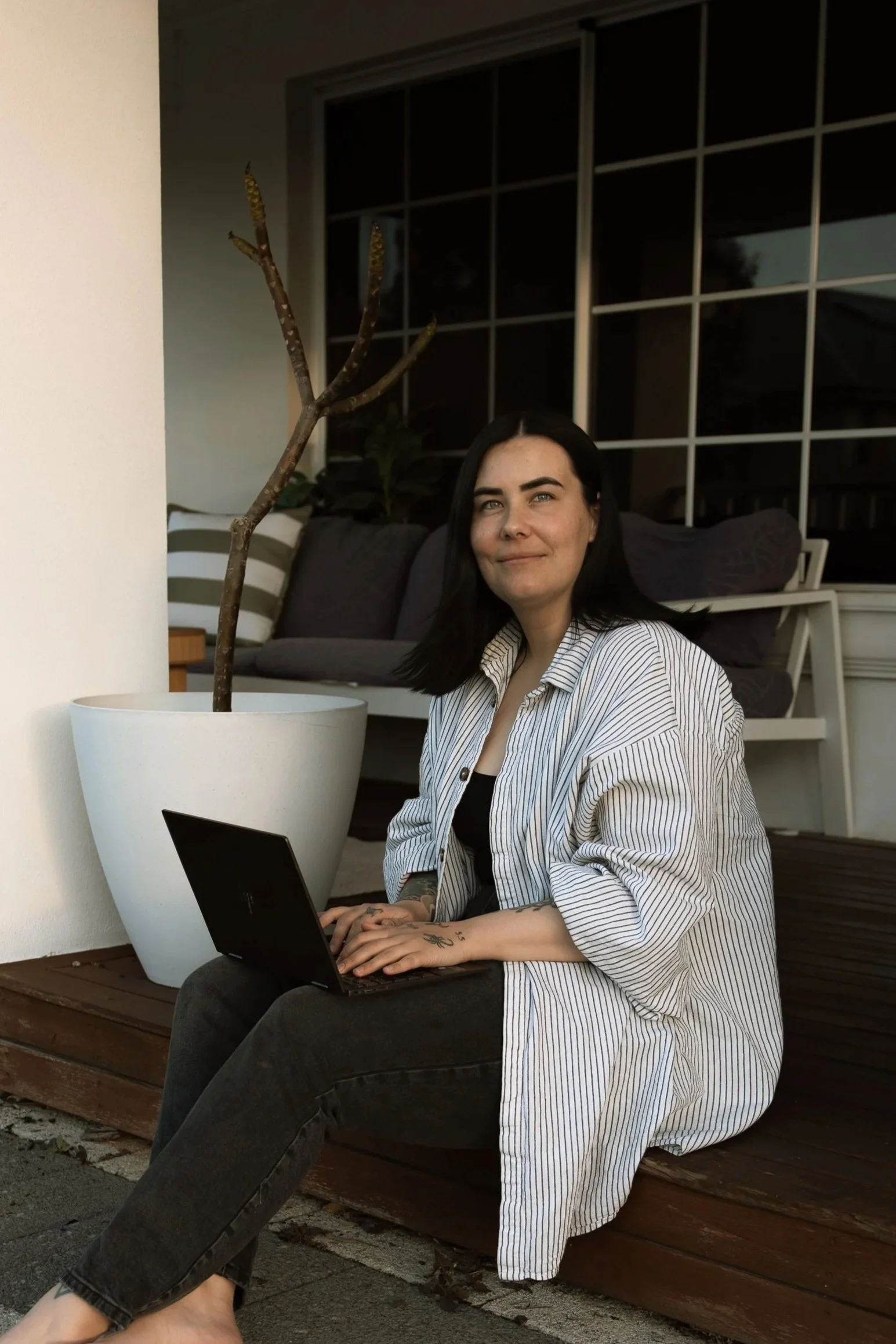 A woman with dark hair and a striped shirt sitting on a wooden platform outside, working on a laptop next to a large potted plant, with a porch and large window in the background.