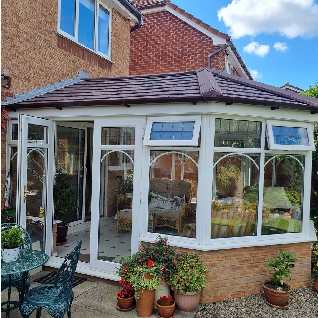A brick house with a conservatory extension featuring large glass windows and white frames, surrounded by potted plants and outdoor furniture.