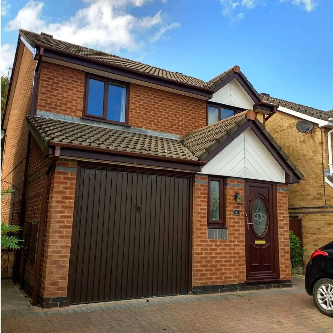 A two-story brick house with a brown garage door, a dark red front door with an oval glass window, and a small window next to the front door. The house has a tiled roof with multiple gables, and there is a small tree or bush on the left side. The sky is partly cloudy with patches of blue.