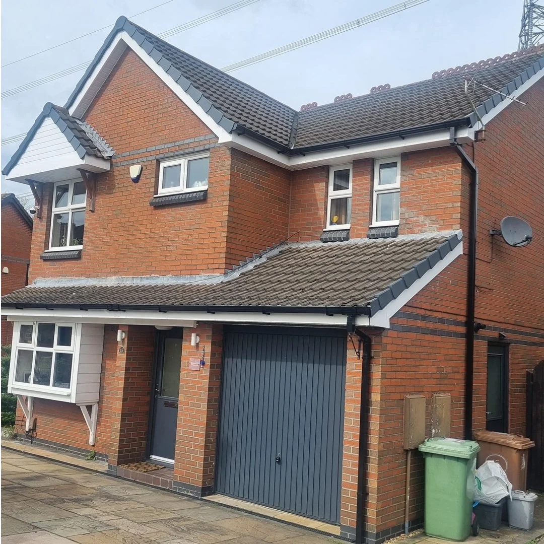 A two-story brick house with a tiled roof, white window frames, and a grey garage door. Multiple trash bins are positioned near the side of the house.