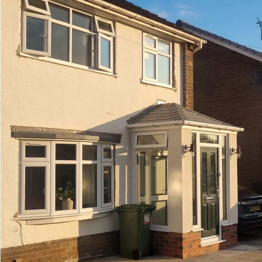 Front of a two-story house with cream-colored stucco exterior, brown brick foundation, a black front door with glass panel, and multiple white-framed windows including a bay window. Sunlight casts shadows on the house.