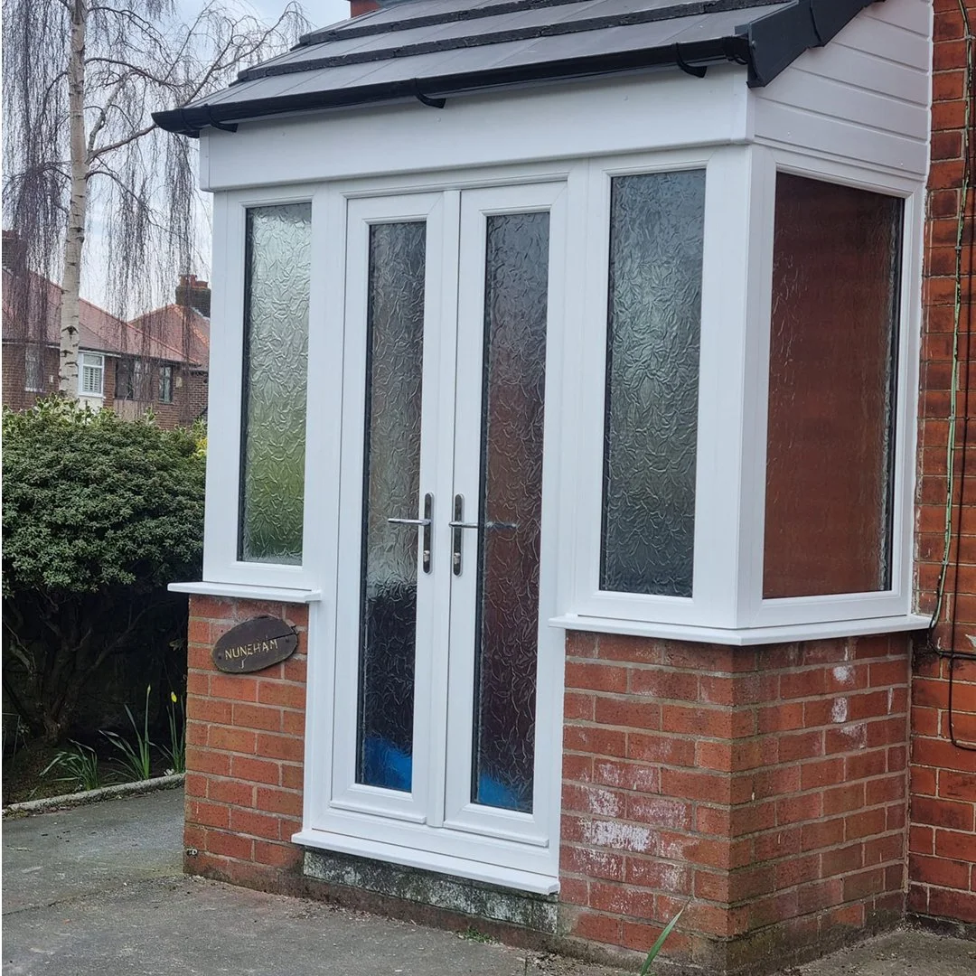 Brick front porch with white bay window and double glass doors, textured glass panels, small porch plaque reading 'Nuneham', in a suburban neighborhood with trees and nearby houses.