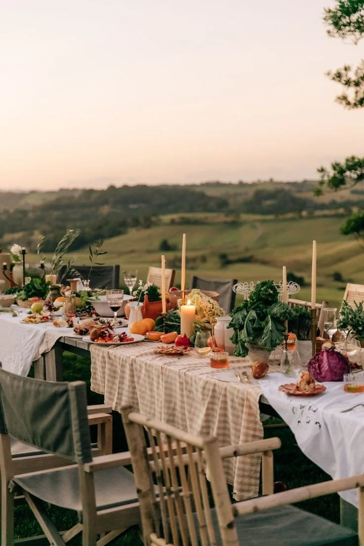Table de dîner en plein air avec des bougies, des fruits, des légumes, de la vaisselle, des verres à vin et des décorations, avec un paysage rural en arrière-plan au coucher du soleil.