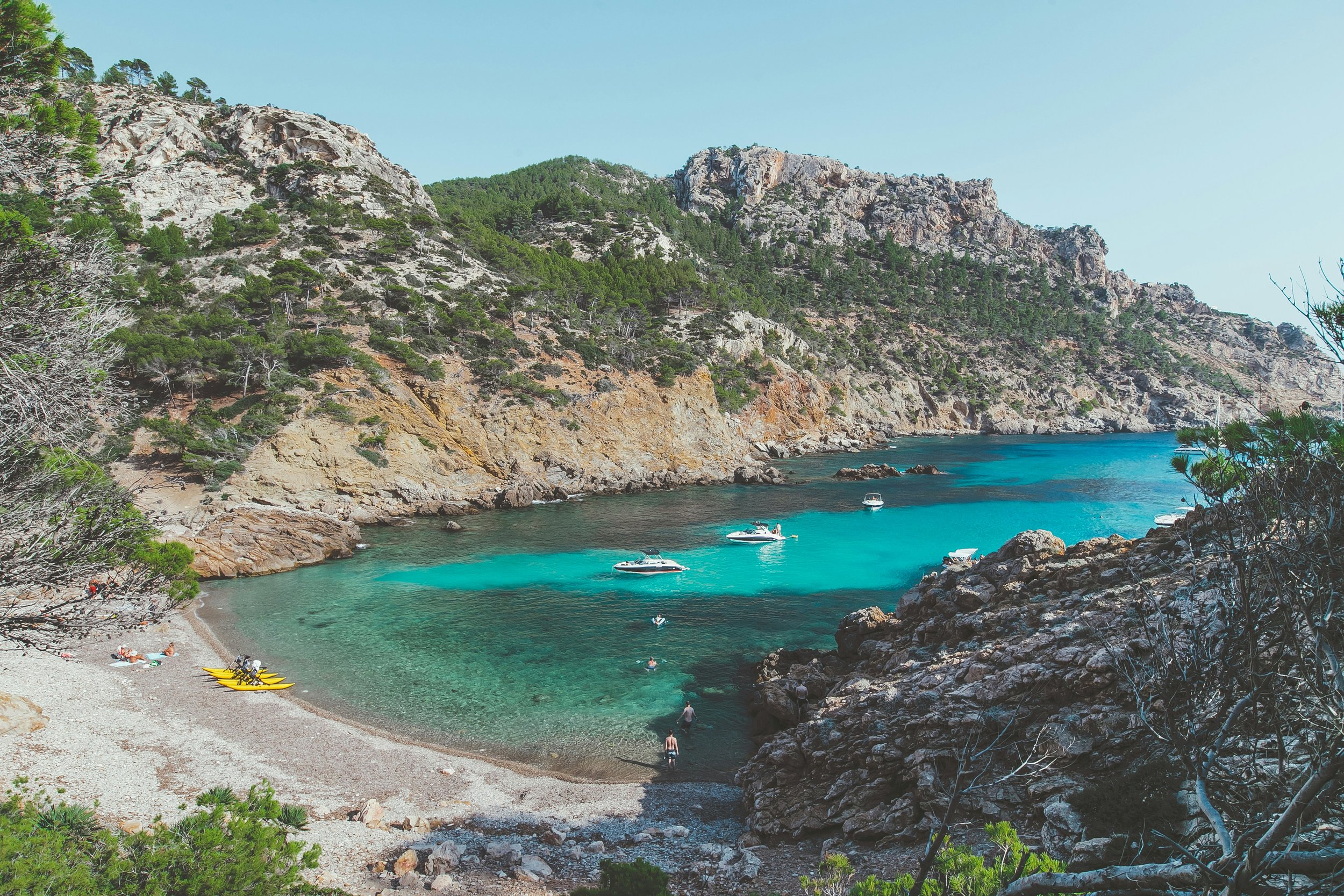 Plage rocheuse avec eaux turquoise, bateaux et personnes nageant, entourée de collines verdoyantes et rocheuses sous un ciel clair.