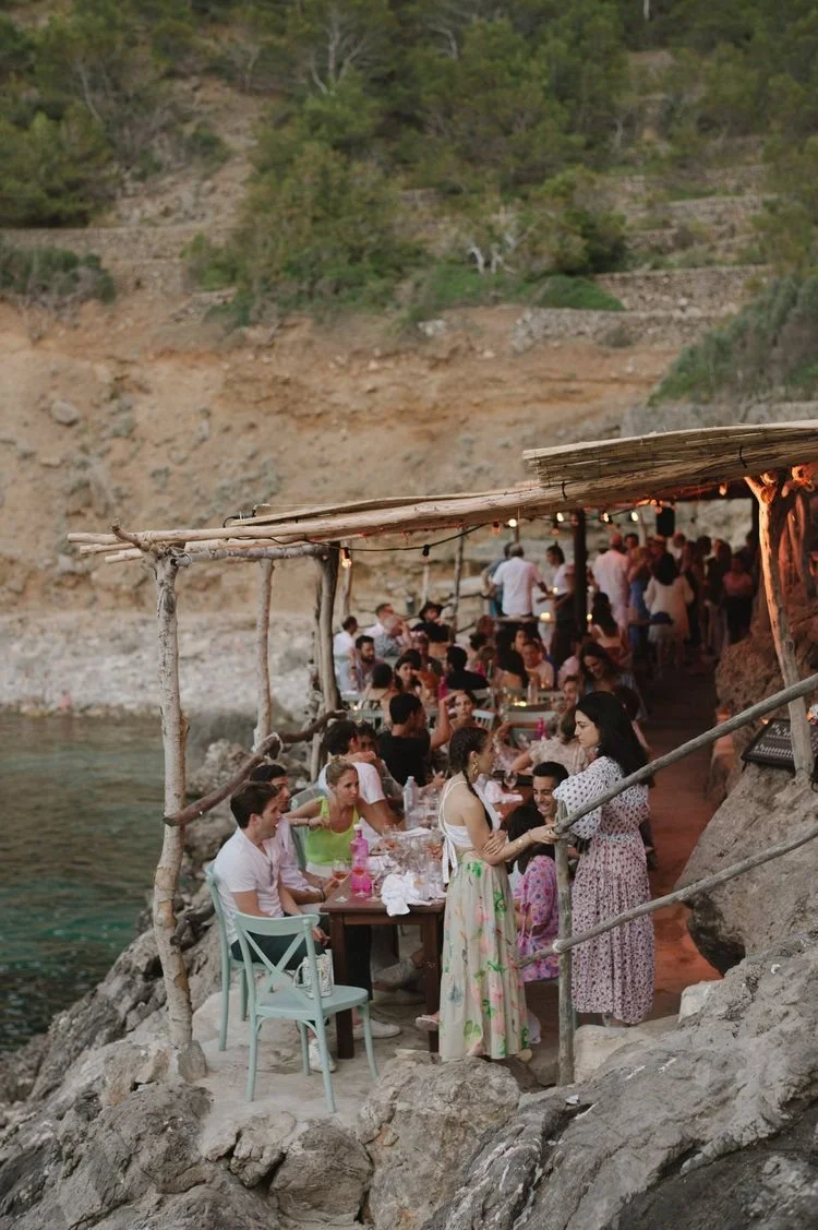 Un groupe de personnes assises et se mêlant à un dîner en plein air sous un abri en bois sur la côte, entouré de rochers et de verdure.