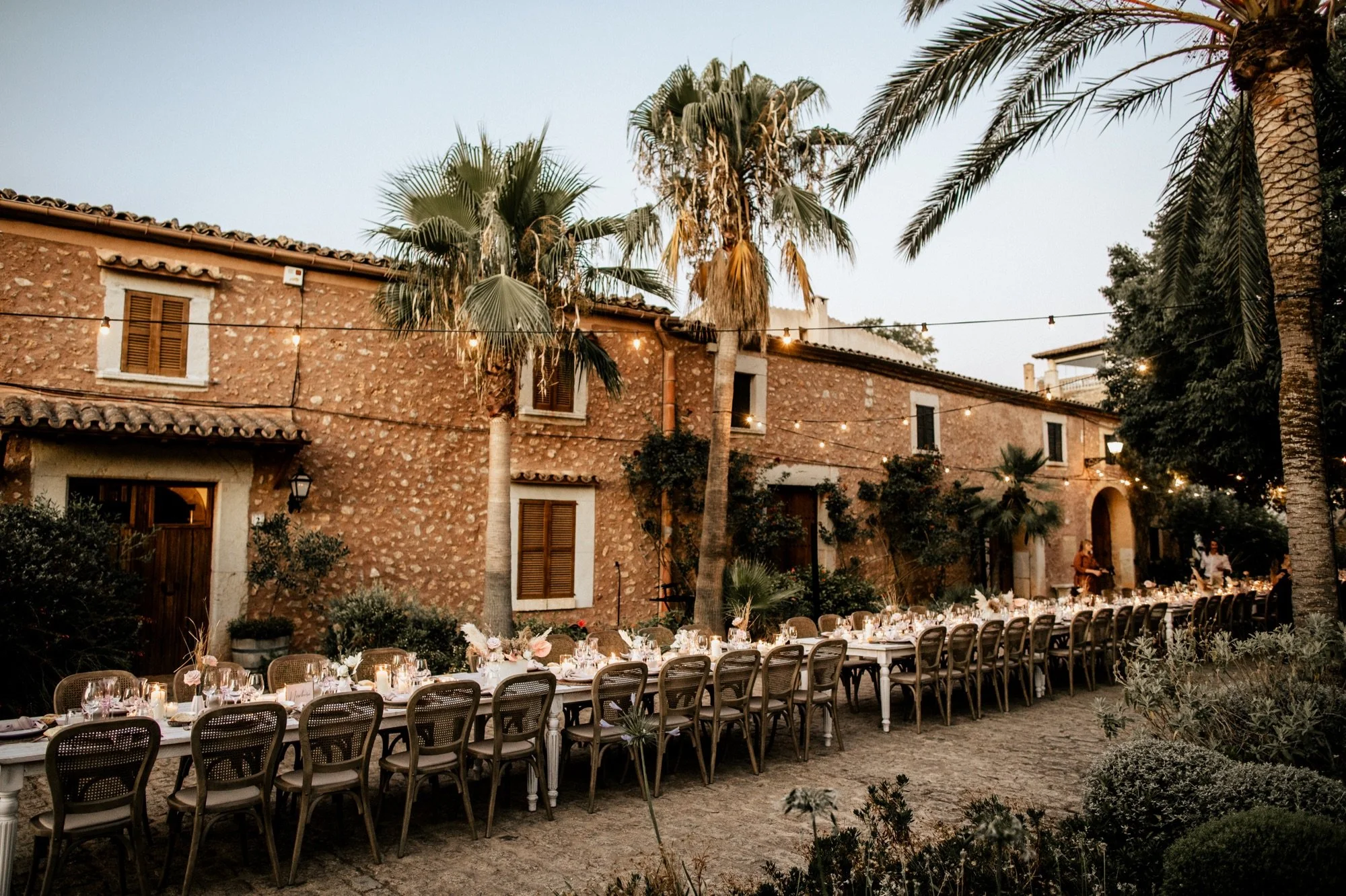 Longue table élégante dressée pour un dîner en plein air, entourée de chaises, avec des bougies et des fleurs, dans un jardin avec des palmiers et une maison en pierre avec des fenêtres à volets en bois, éclairée par des guirlandes lumineuses, le soir.