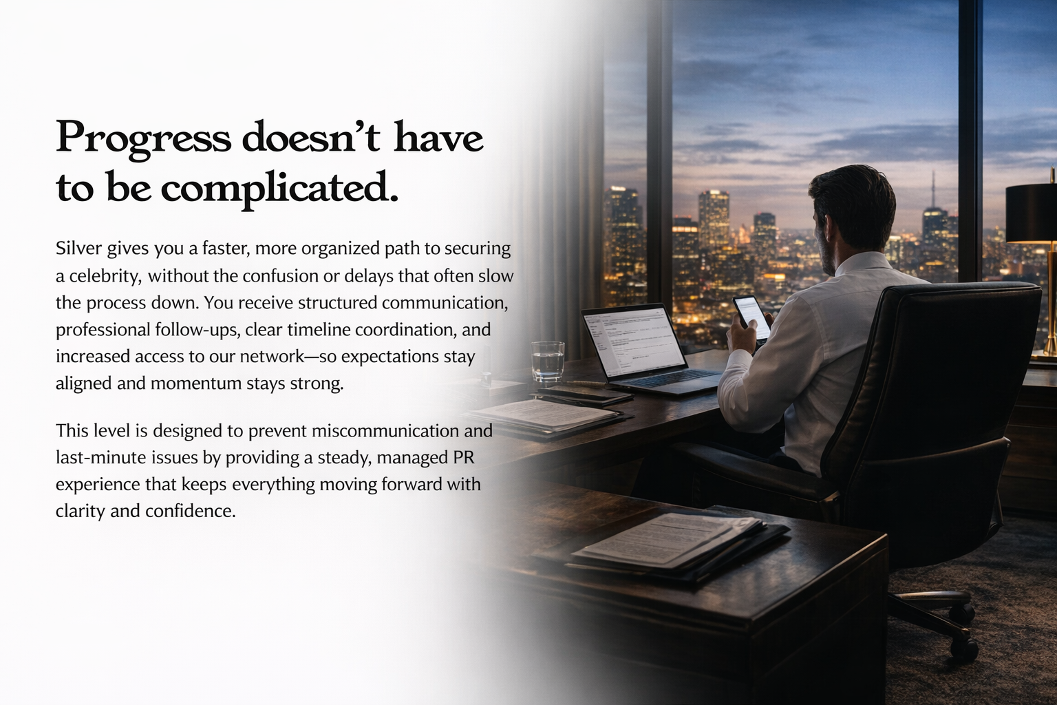 A man sitting at a desk in an office with a city skyline view at dusk, working on a laptop and looking at his phone.
