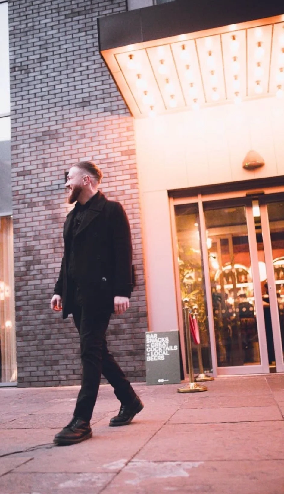 A man with a beard and styled hair, dressed in black, walking past a restaurant or bar with bright lighting and a glass door. There is a sign outside listing drinks and snacks.