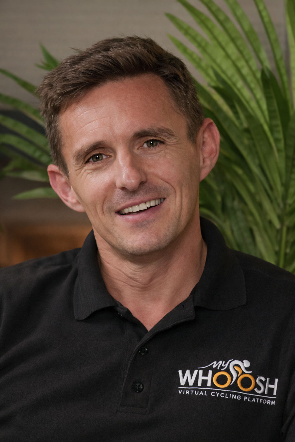 A smiling man wearing a black polo shirt with a logo that reads "my WHOOSH VIRTUAL CYCLING PLATFORM". He is sitting indoors with green plants in the background.