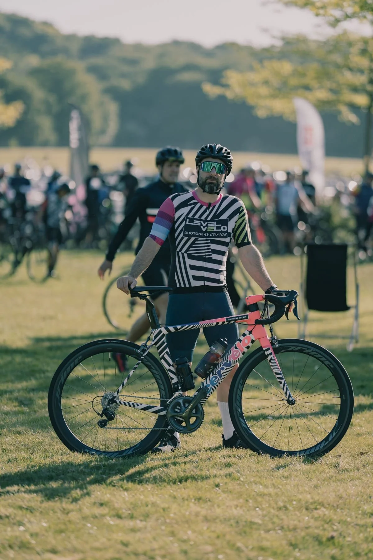 A man in cycling gear and a helmet standing with a pink and black road bike on a grassy field during a cycling event, with other cyclists and event tents in the background.