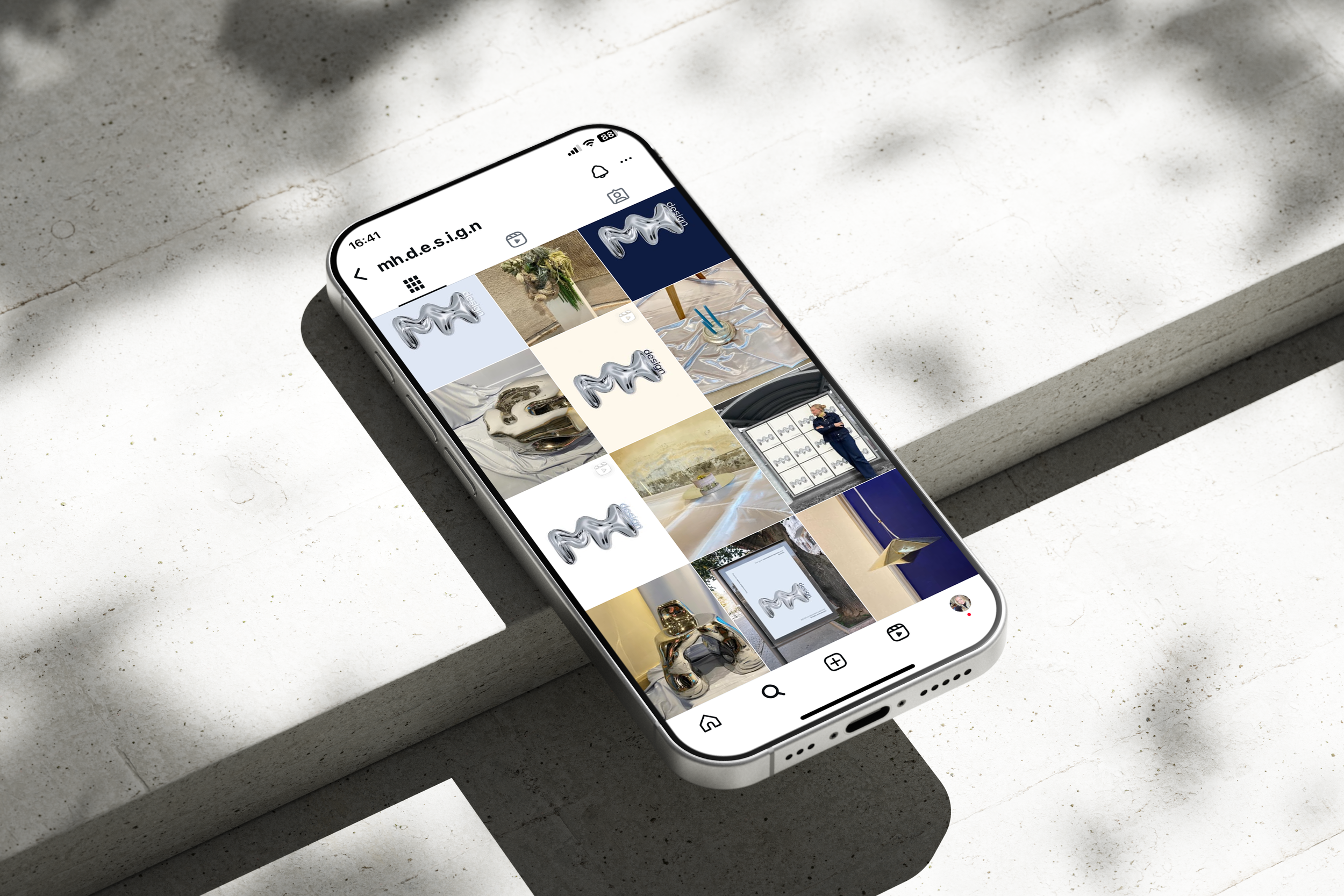 En hvit smarttelefon ligger på en lysegrå betongflate, og viser en Instagram-profil med ulike bilder av dekorative objekter og kunst. Skjermen er delvis i skygge.