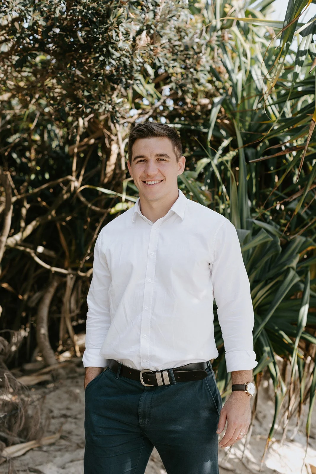 A young man standing outdoors on a sandy surface with lush green foliage behind him, smiling and wearing a white long-sleeve shirt, dark pants, a watch, and a belt.