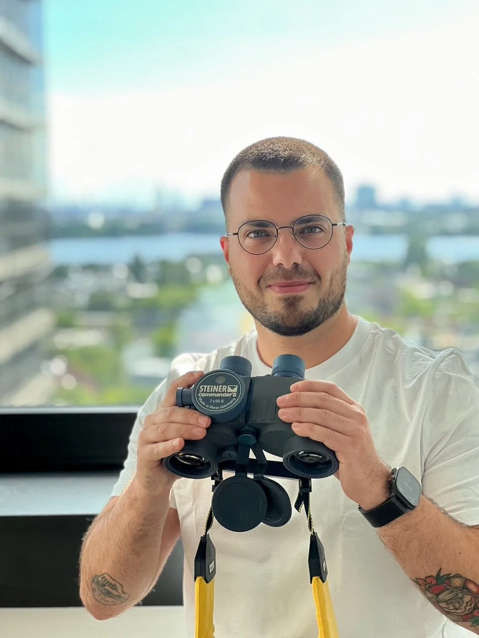 Ein Mann mit Brille hält ein schwarzes Fernglas mit gelben Bändern vor sich, steht vor einem Fenster mit Stadt- und Wasserblick, trägt ein weißes T-Shirt und eine Watch am linken Handgelenk.
