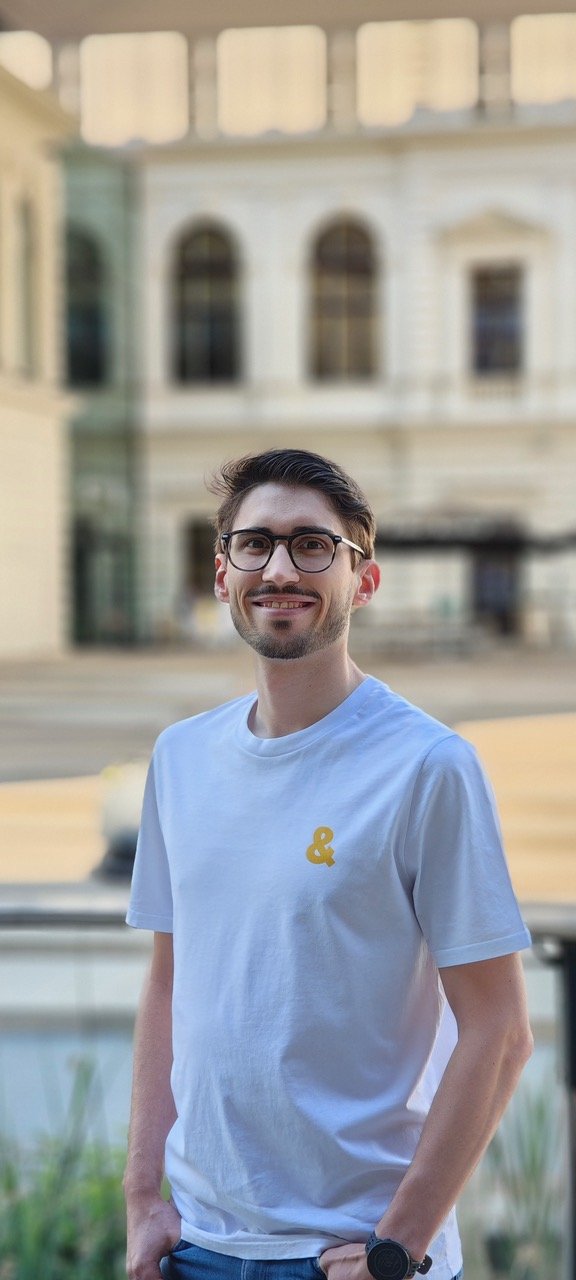 Ein junger Mann mit Brille, weißen T-Shirt mit gelbem Logo auf der Brust, steht draußen vor einem historischen Gebäude.