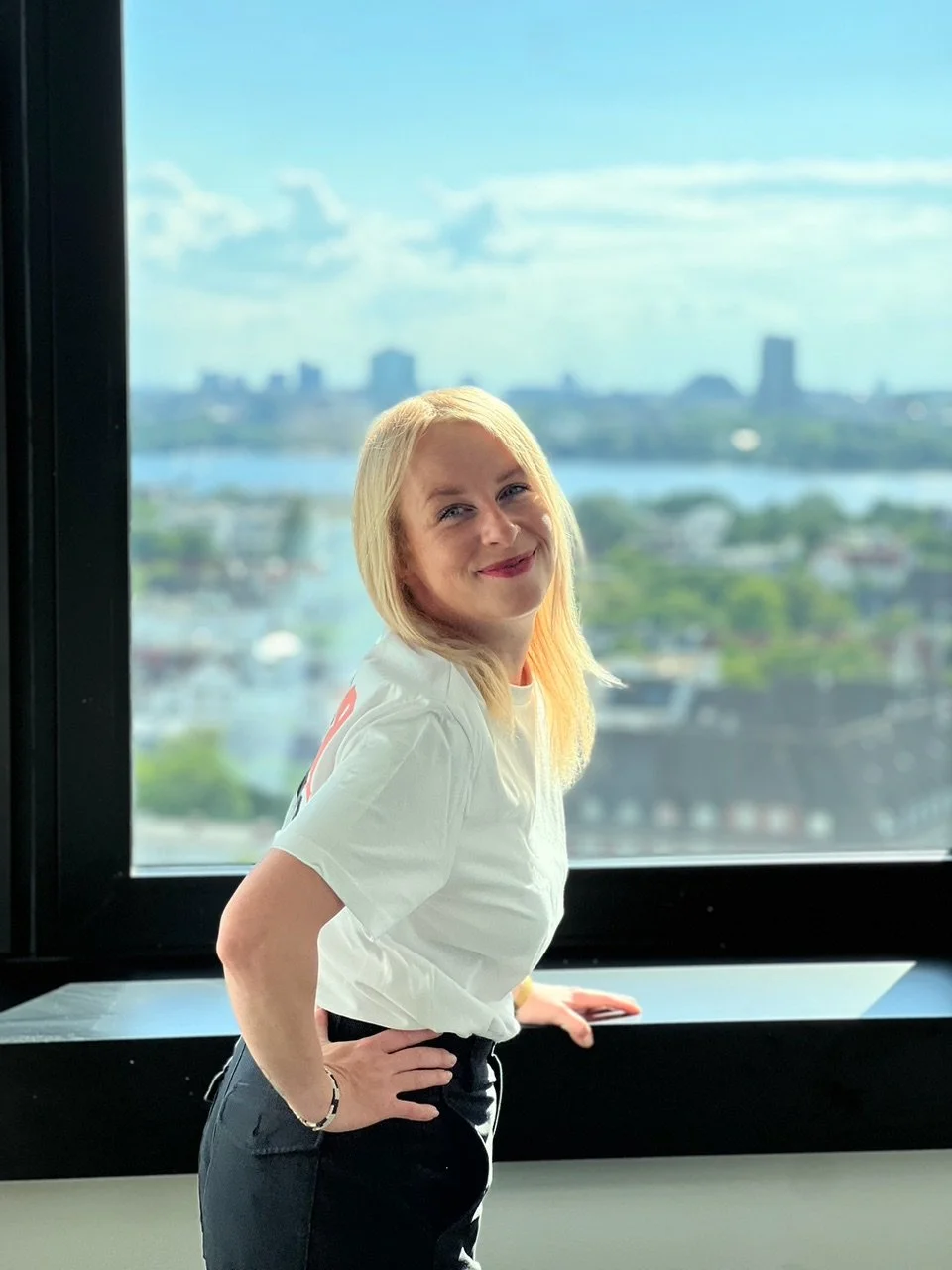 Eine Frau mit blonden Haaren steht vor einem großen Fenster mit Stadt- und Flussblick, lächelt und trägt ein weißes T-Shirt und dunkle Hosen.