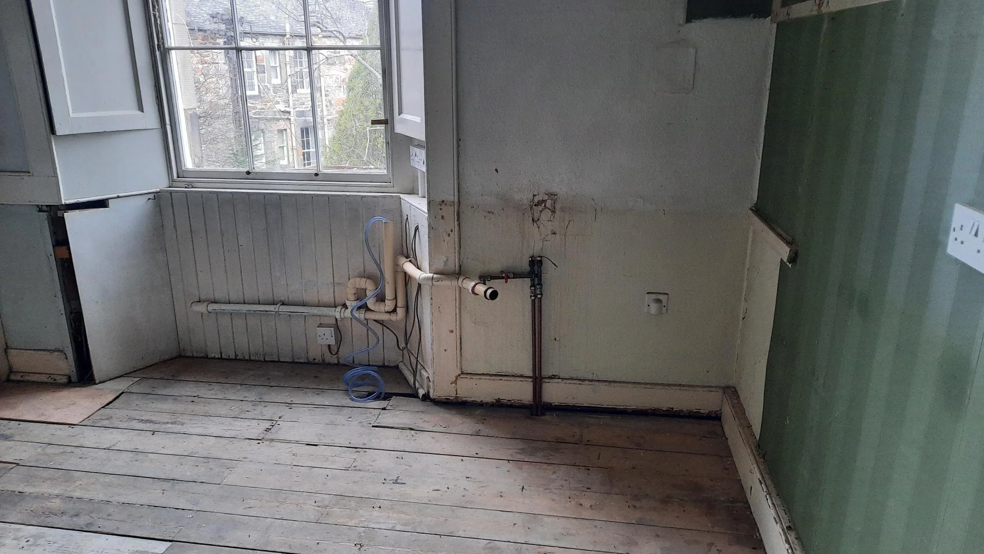 An unfinished kitchen with a window showing an outside apartment building, with exposed plumbing and electrical outlets, and partially removed flooring and wall panels.