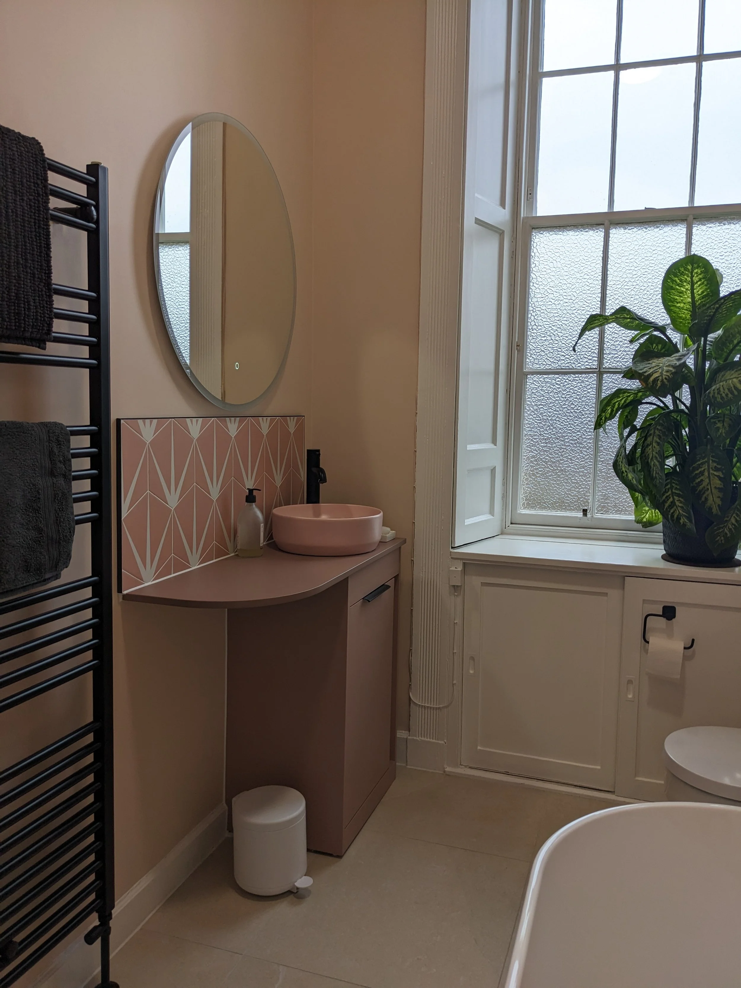 A bathroom with a pink vanity, round pink sink, large window with a potted plant, mirror, black towel rack, and a small white trash can.