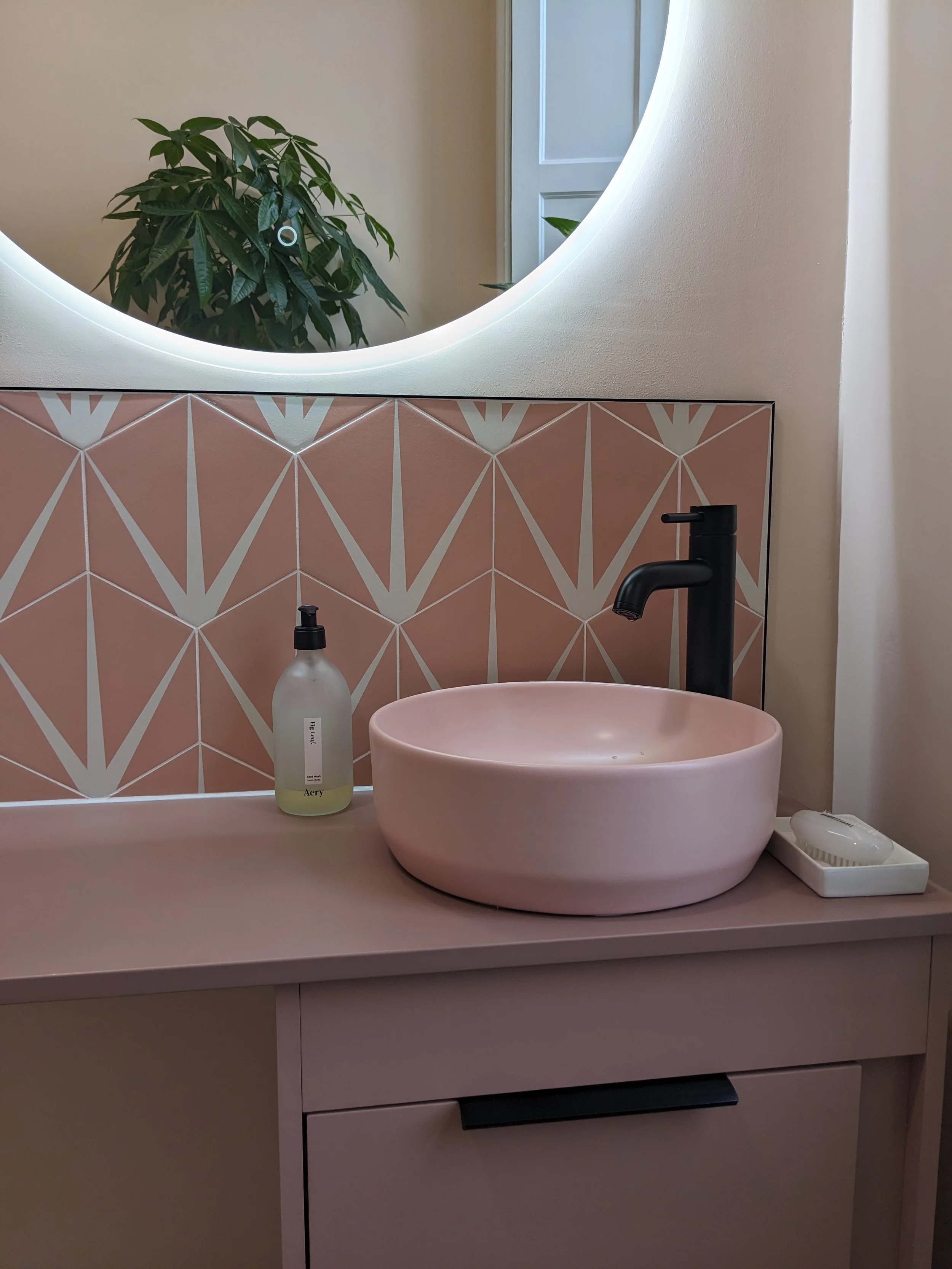 A bathroom sink area with a pink vessel sink, black faucet, a soap dispenser, and a mirror with circular lighting. A plant is reflected in the mirror, and the backsplash has geometric patterned tiles in pink and white.