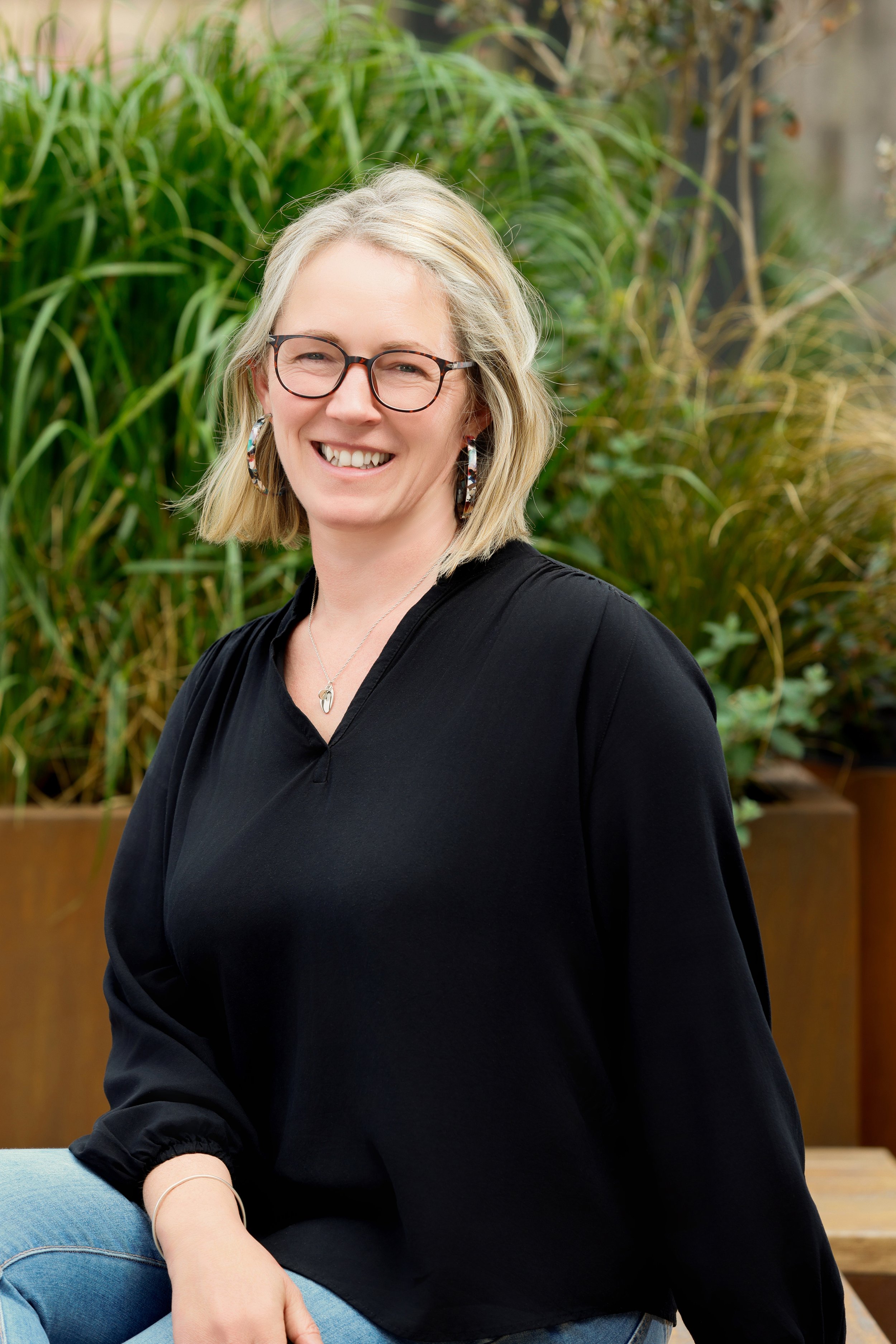 A smiling woman with blonde hair, wearing glasses, earrings, and a black top, sitting outdoors with green plants in the background.