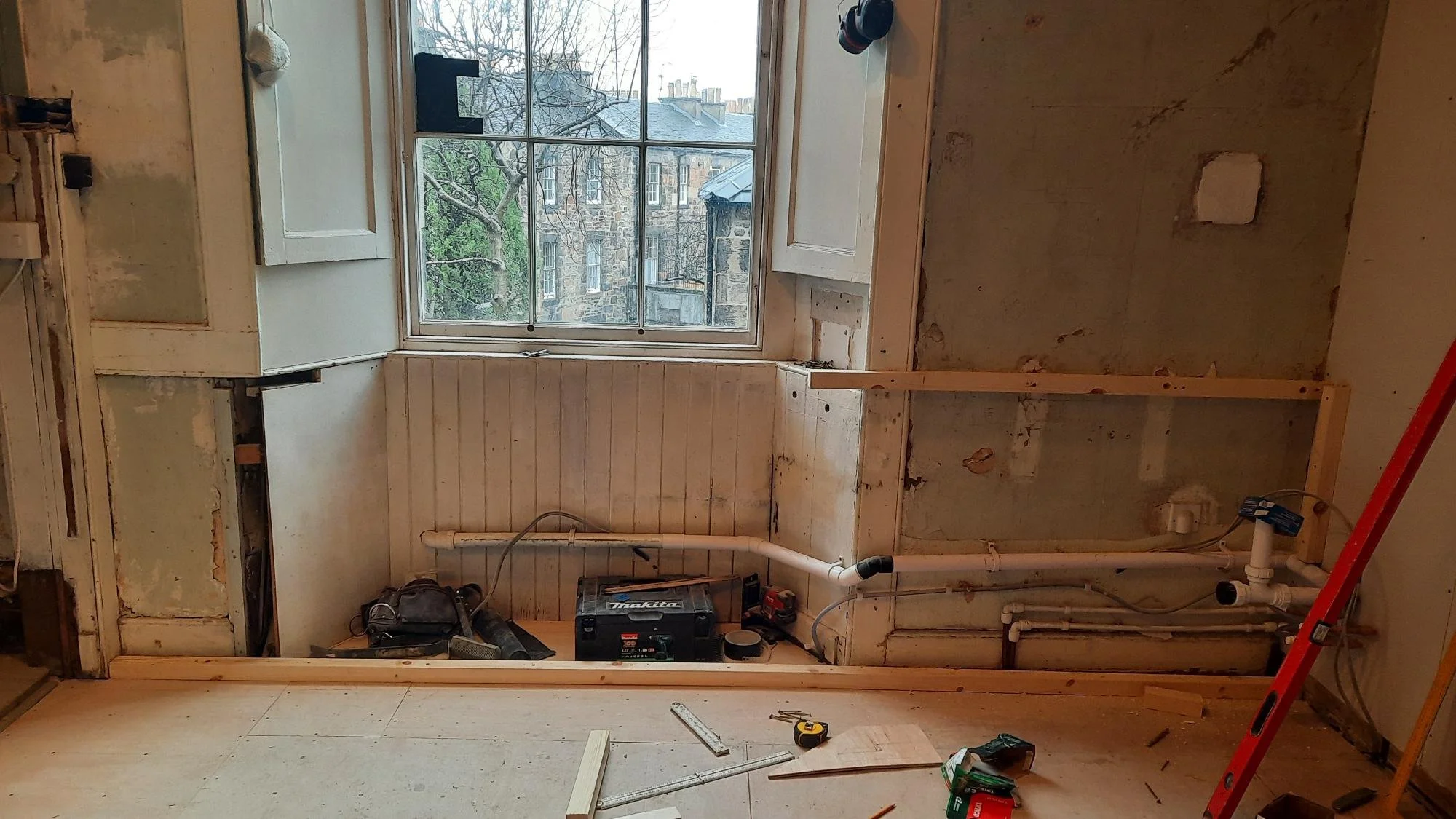 Room under renovation with exposed plumbing and construction tools, window showing neighboring buildings, and unfinished walls