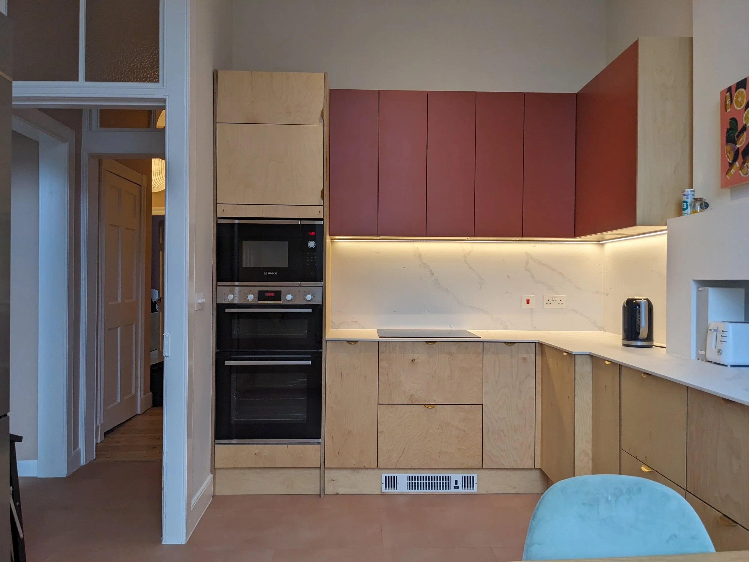 Modern kitchen with beige wood cabinets, red upper cabinets, marble backsplash, built-in convection oven and microwave, black kettle, white toaster, and a blue upholstered chair in the foreground.