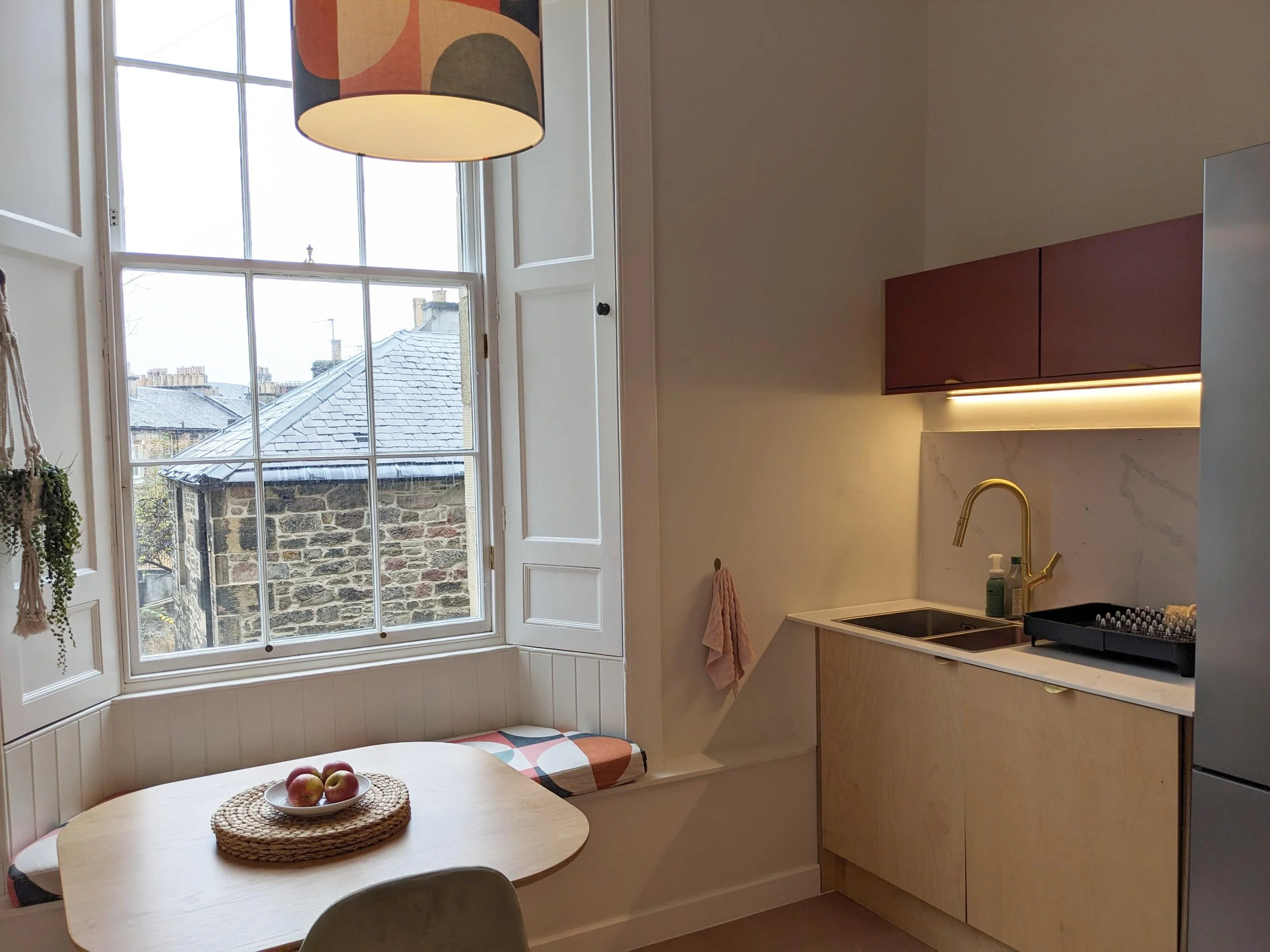 A kitchen with a window seat, a round table with apples, a hanging lamp, and a compact kitchen with a sink, gold faucet, pink cabinets, and a marble backsplash.